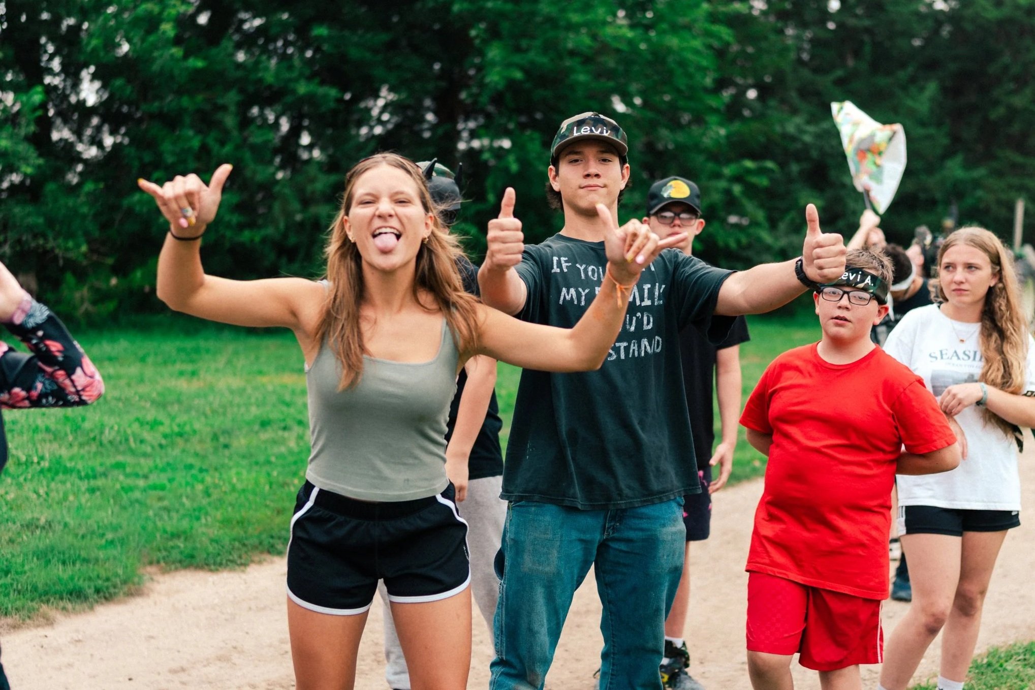 Group of teenagers outdoors, some smiling and making gestures, with greenery and trees in the background.