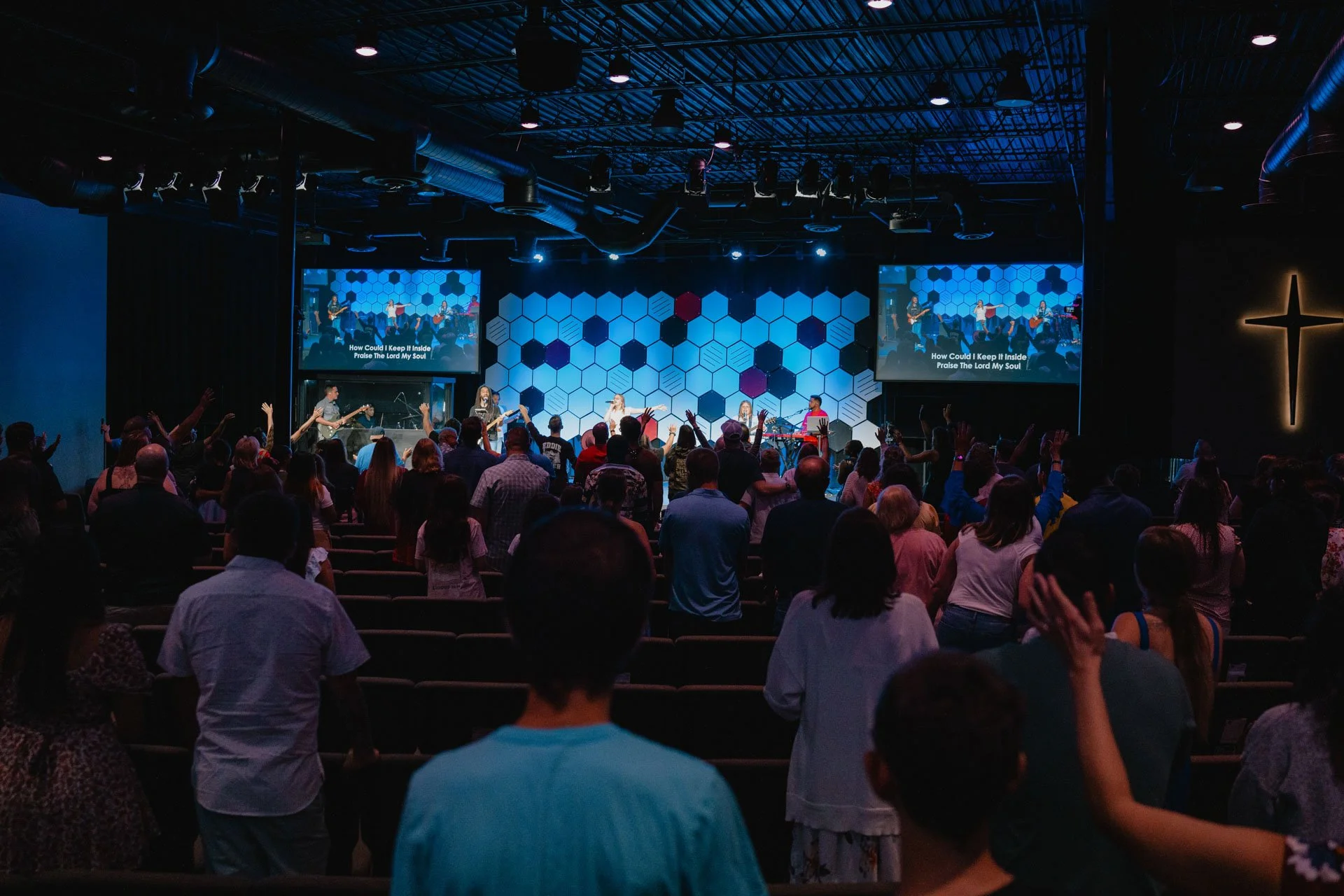 A church congregation standing and engaging in worship during a service, with a band performing on stage and anthem lyrics displayed on two large screens.