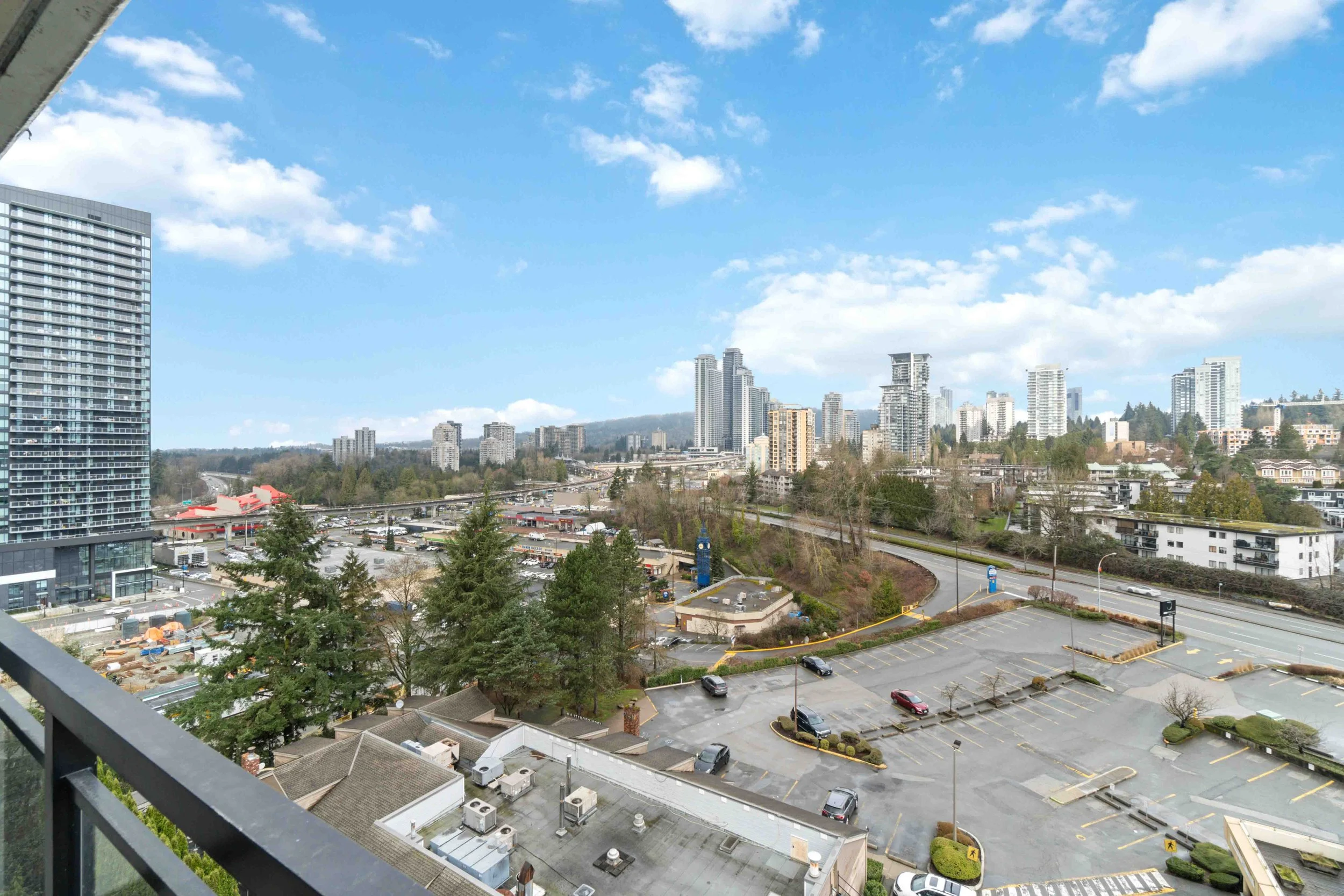 Cityscape view from a high-rise balcony showing a parking lot, mid-rise buildings, and a downtown skyline under partly cloudy skies.