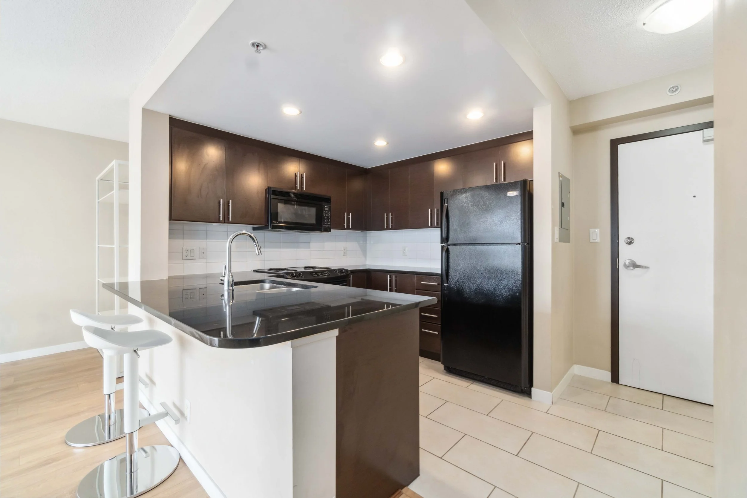 Modern kitchen with dark wood cabinets, black refrigerator, microwave, stove, and white tile backsplash, with a breakfast bar and white barstools.