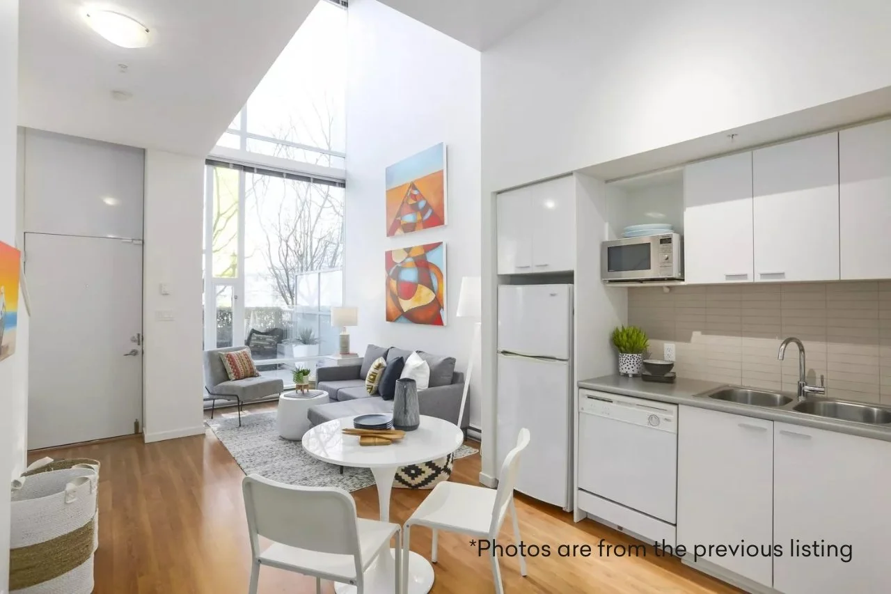 Modern open-concept living room and kitchen with white walls, large windows, gray sofa, white round dining table, and art on the wall.