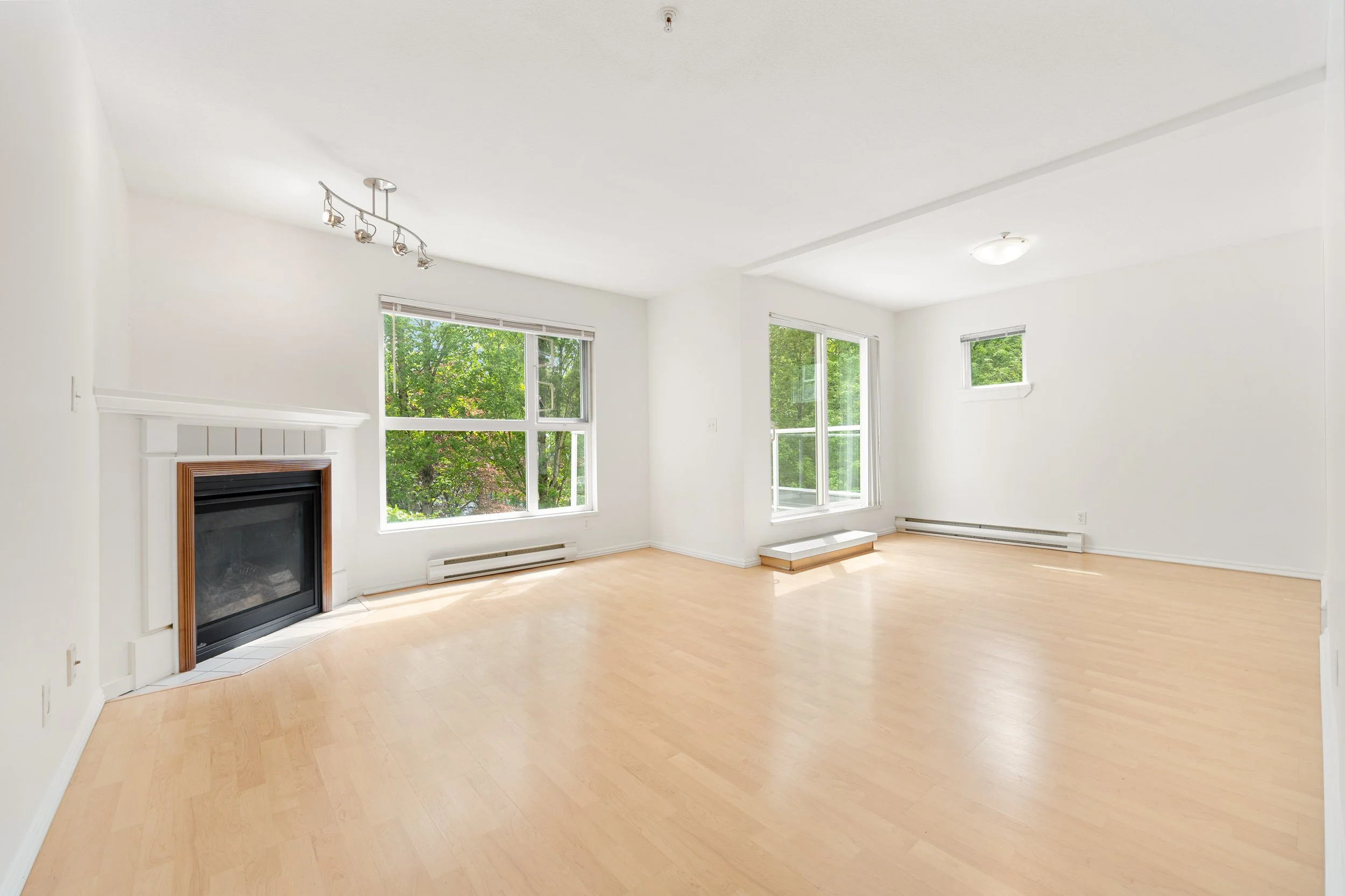 Empty living room with large windows, a fireplace, light wood flooring, and white walls.