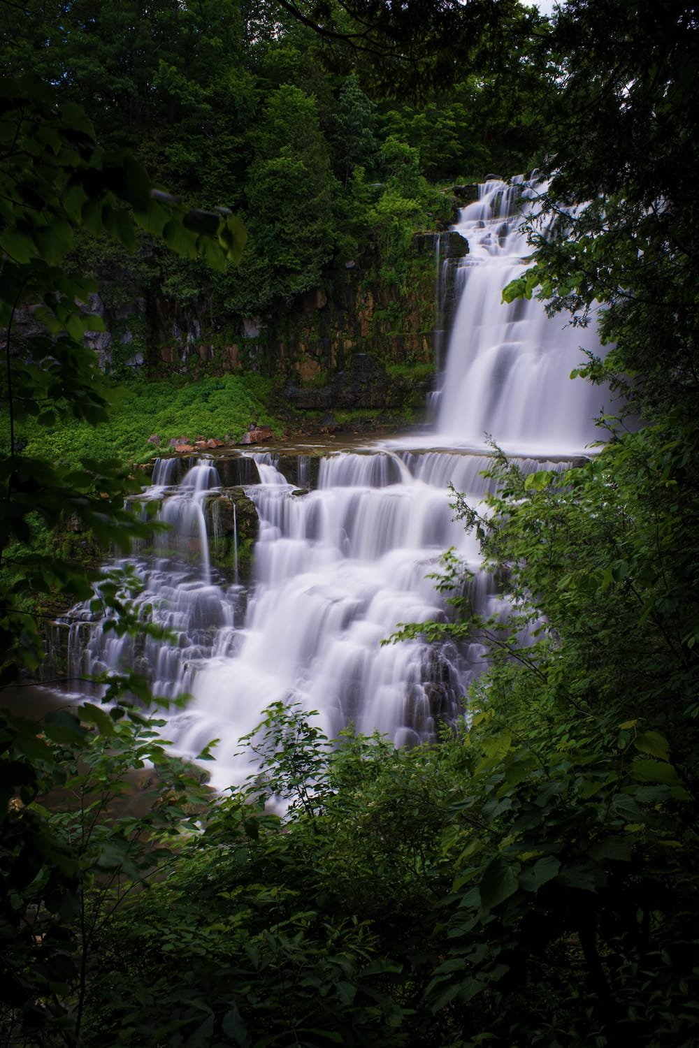 A multi-tiered waterfall flowing through a lush, green forest with dense foliage on all sides.
