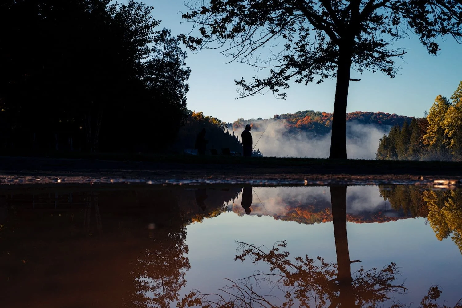 Two people fishing by a calm lake at dusk, with mist rising from the water and trees with autumn foliage in the background. The image is reflected in the still water, creating a mirror effect.