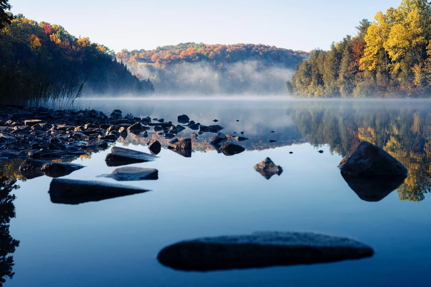 Calm river with rocks near the shore, mist over the water, autumn-colored trees on both sides, and distant hills in the background.