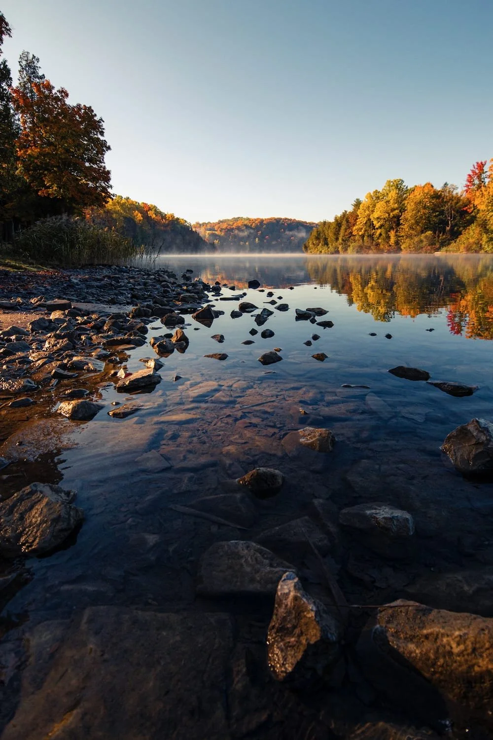 Rocks along the shoreline of a calm river surrounded by trees with autumn foliage, with mist rising over the water and clear blue sky.