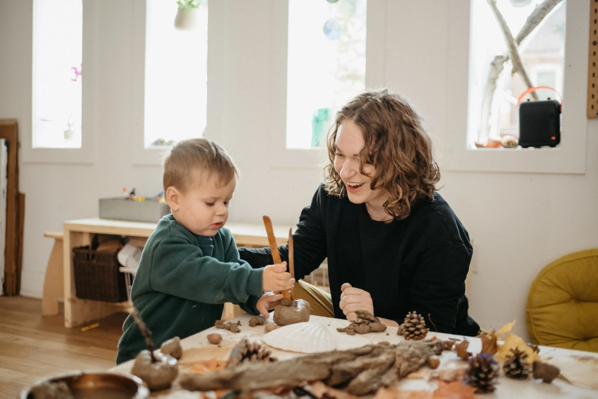 A woman and a child engaged in using clay and natural materials such as pine cones and leaves, in a bright art studio