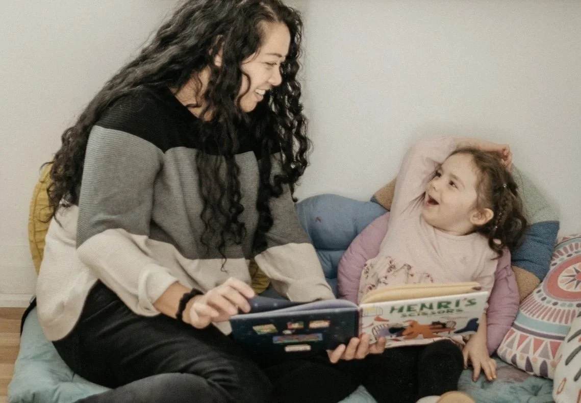 A woman and a young girl sitting together on cushions, smiling and holding open children's books. The woman has long, curly hair and is wearing a striped sweater. The girl has her hand in her hair, smiling joyfully.