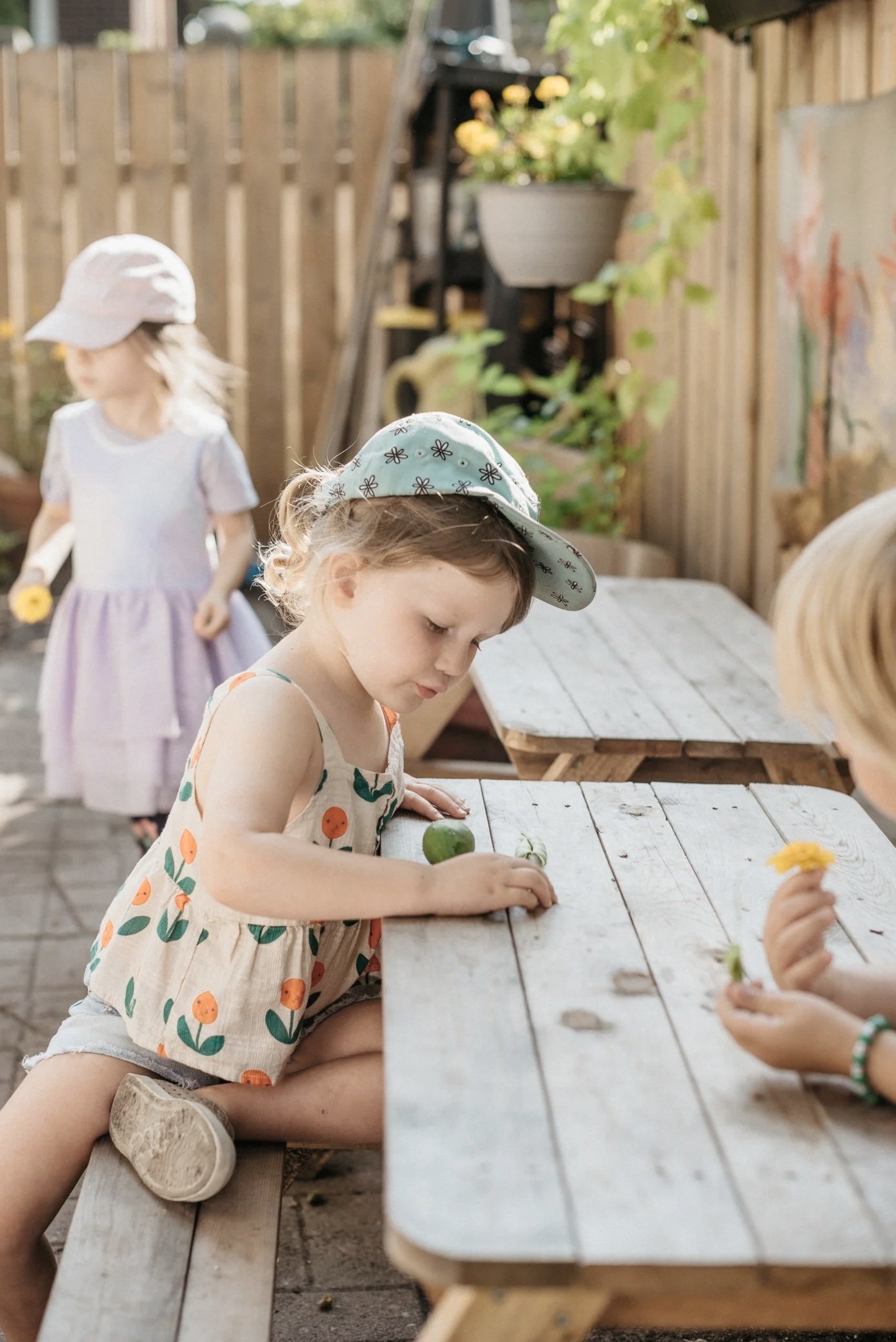 Children sitting at a wooden picnic table outdoors, wearing summer clothes and hats, with a wooden fence and plants in the background. One child is examining a flower or vegetable from the garden.