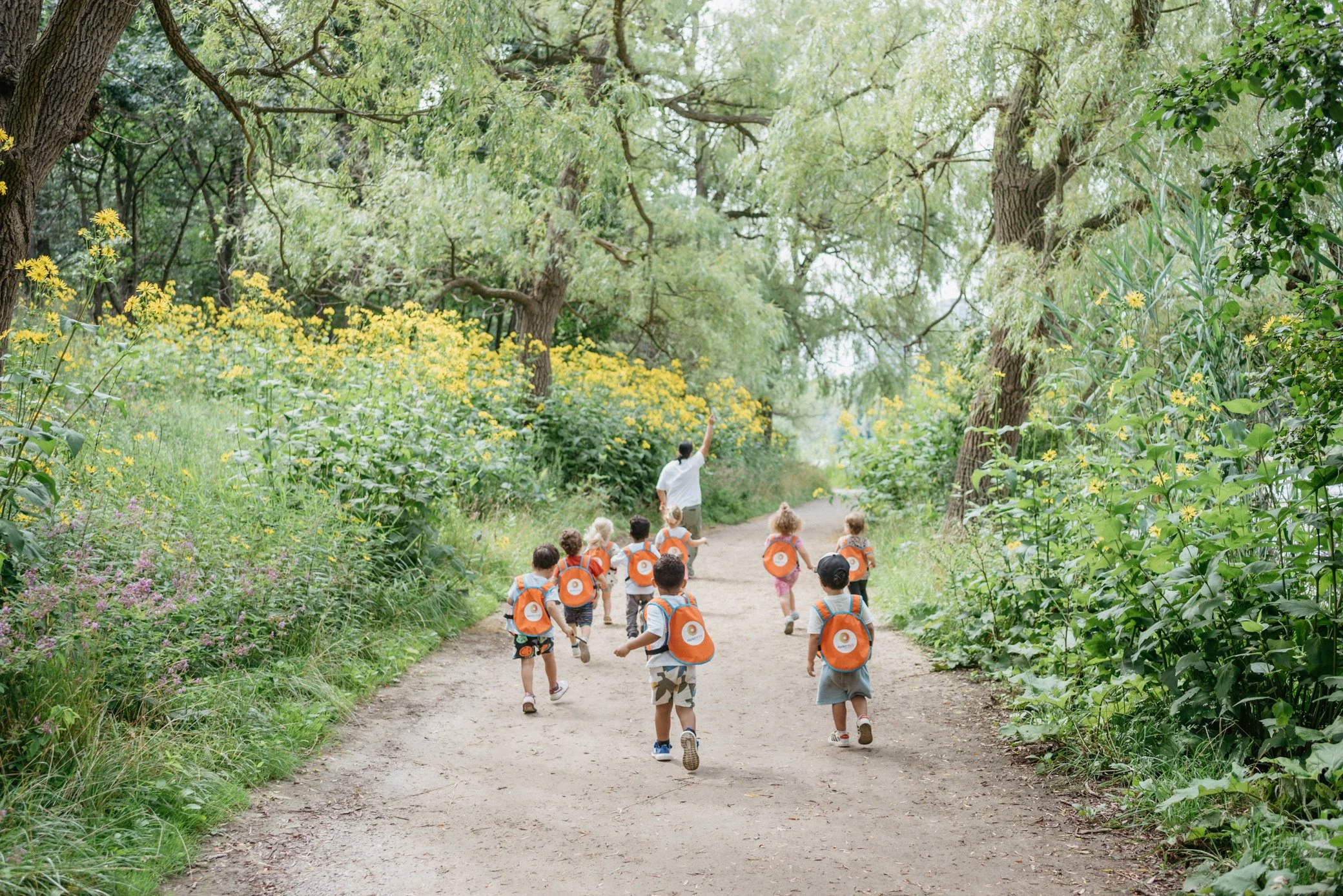A group of young children with orange backpacks walking on a dirt trail through a lush, green forest while a caregiver leads them.
