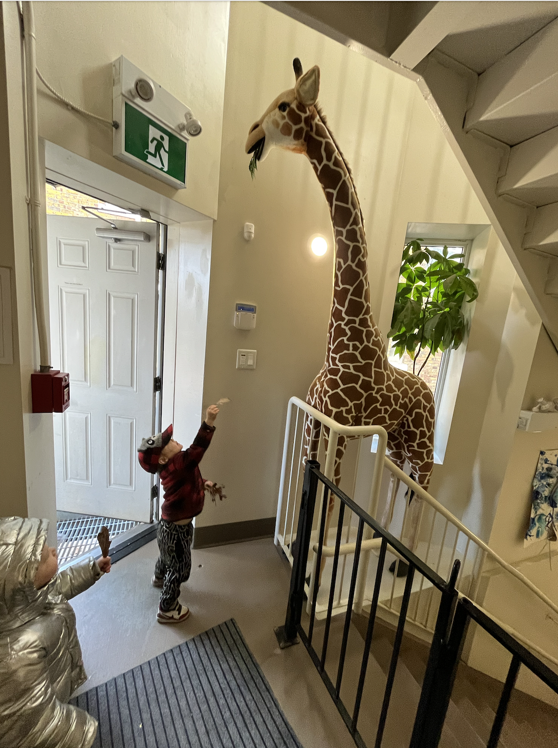 A giant giraffe statue with a leafy branch in its mouth inside a building. Two children, one wearing a metallic jacket and another in a red jacket and hat, appear to be playing or interacting with the giraffe. The setting is a hallway with a door, wi