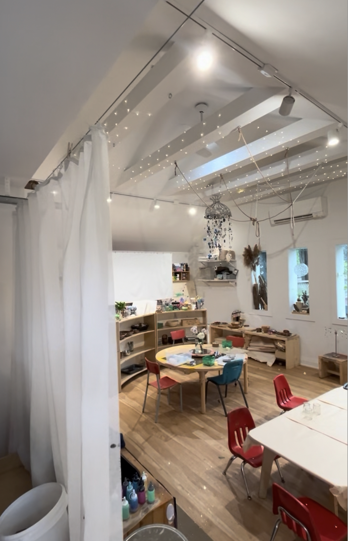 A cozy, well-lit room with ceiling fairy lights, white walls, and wooden floors. There are tables and red chairs, shelves with art supplies, and decorative hanging ornaments on the wall.