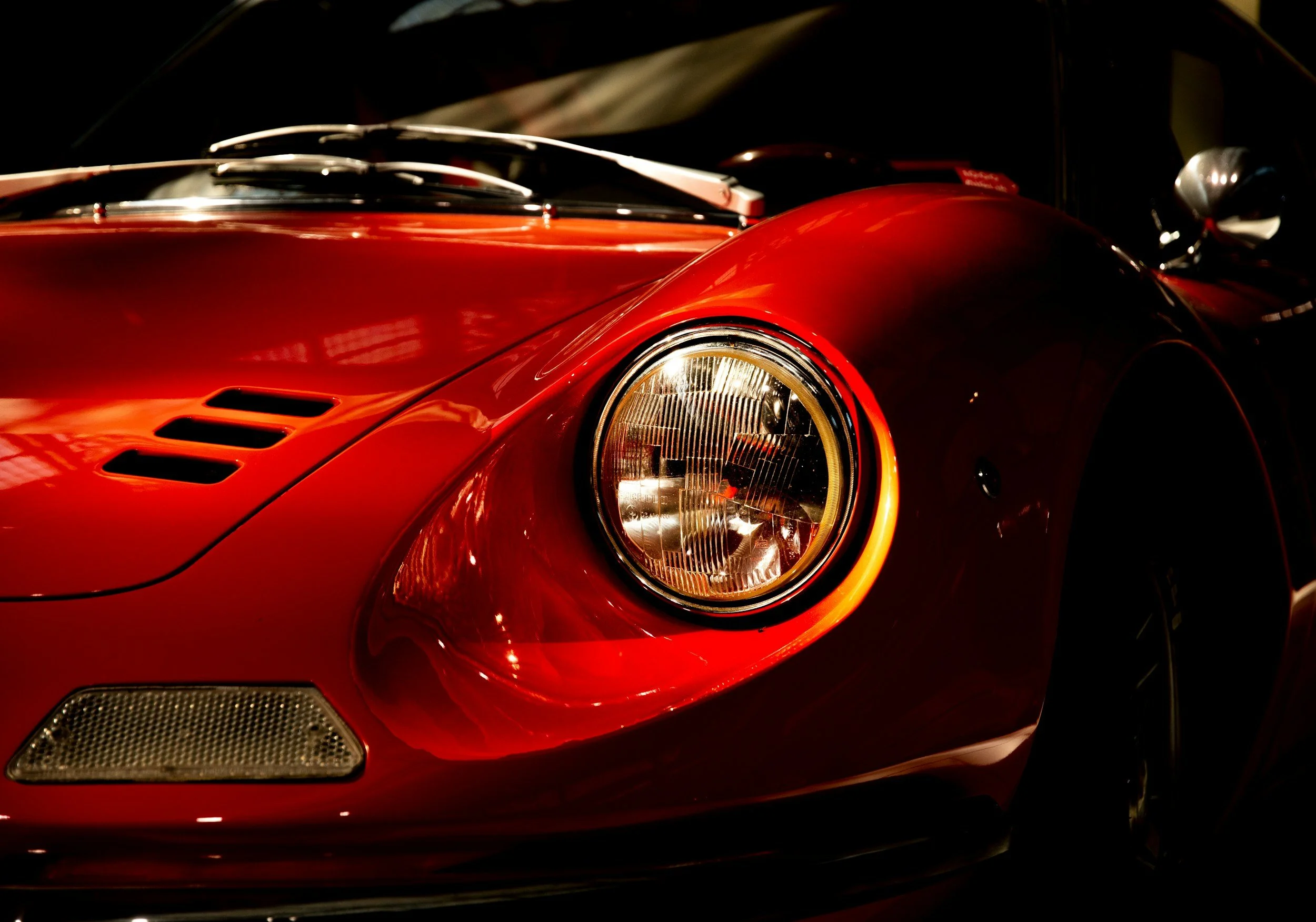 Close-up of the front left side of a red vintage sports car, focusing on the round headlight and part of the hood.