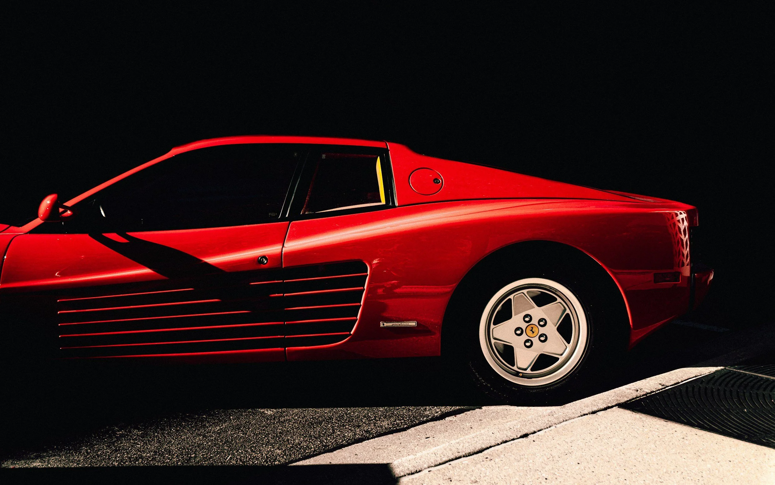 Red classic Ferrari sports car parked on a street at night, with shadows cast on the side.