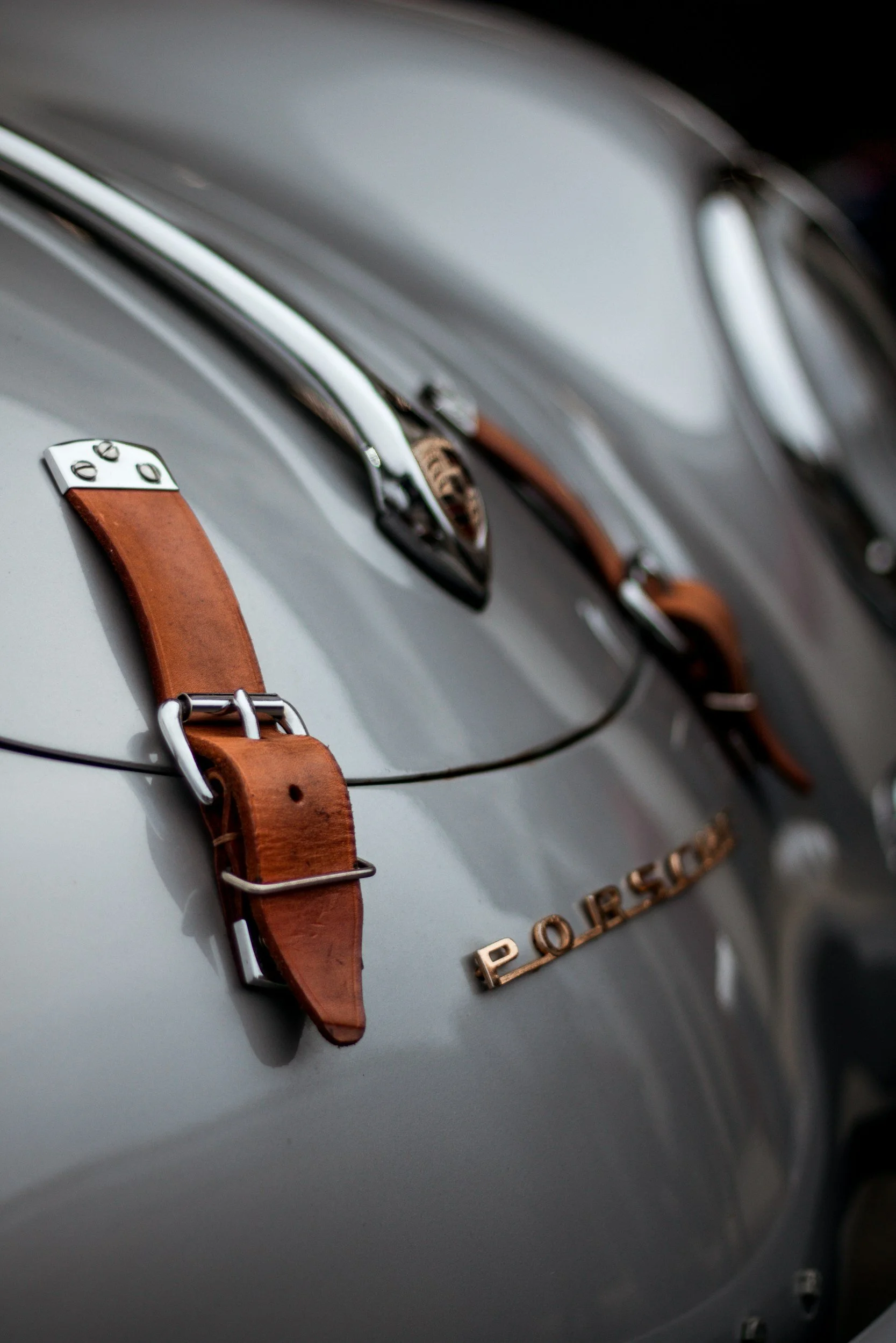 Close-up of a vintage Porsche car badge with leather straps and metal buckles on a gray Porsche vehicle.