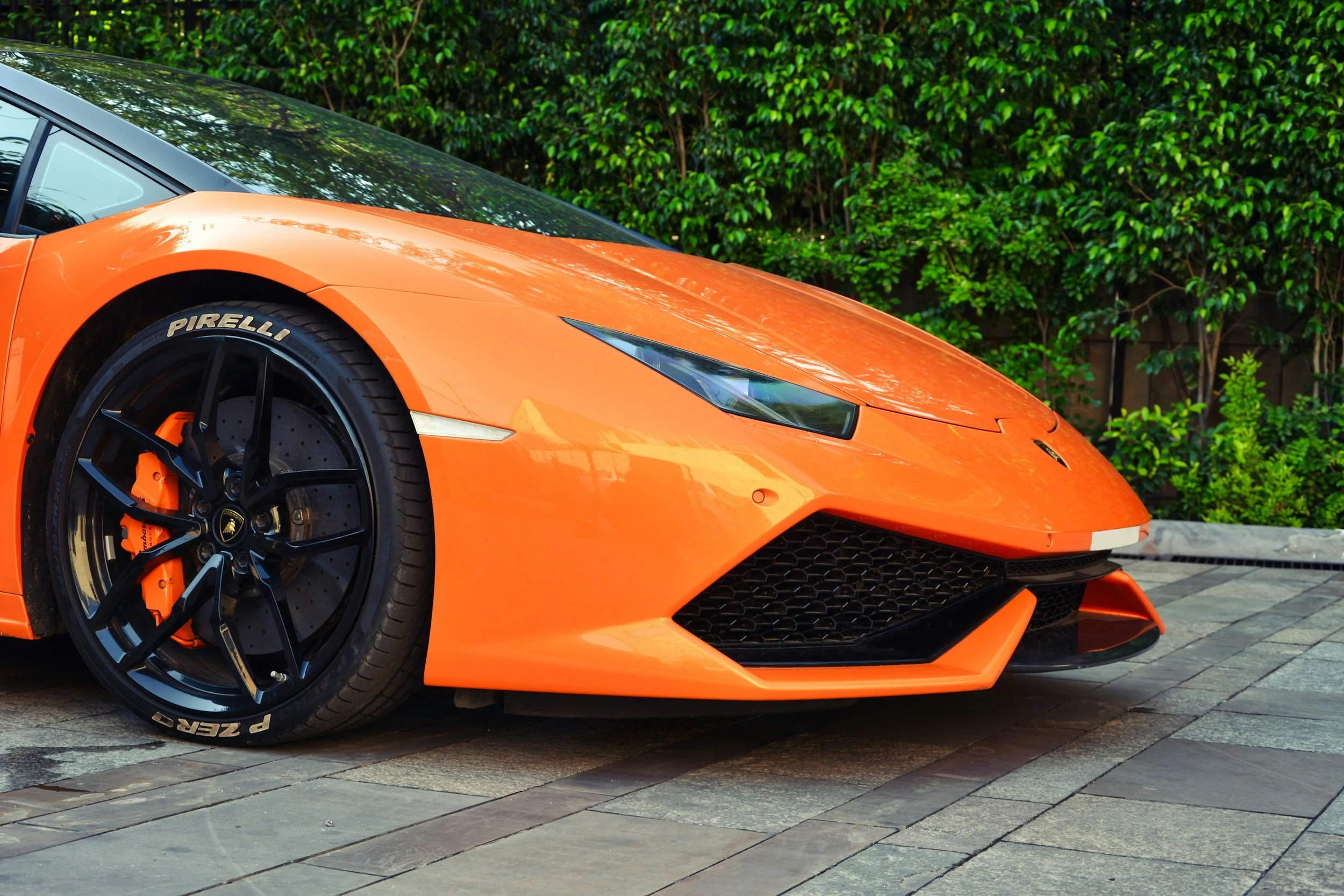 Orange Lamborghini sports car parked on a stone driveway with green hedges in the background.