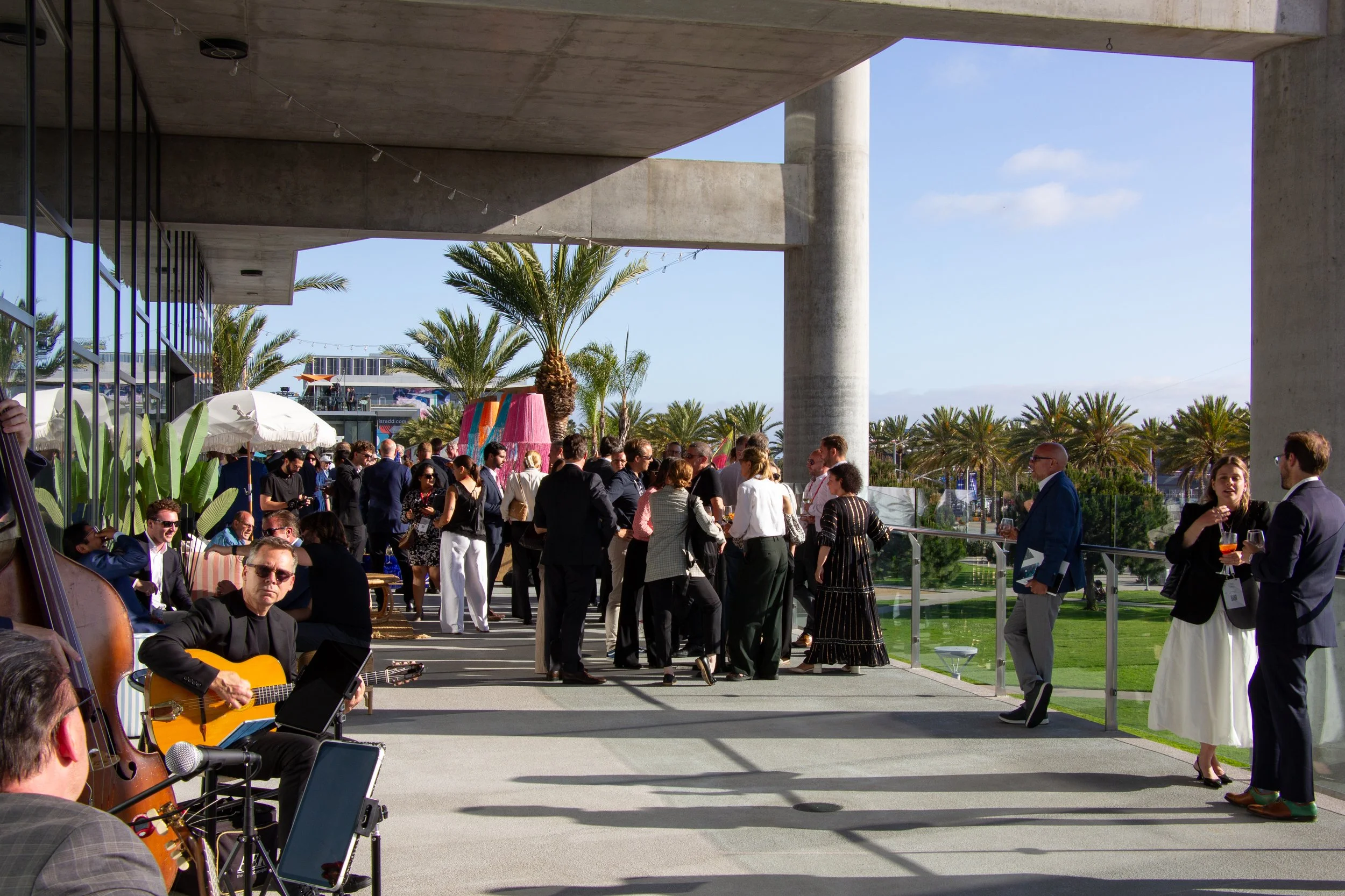 A large group gathered on The Lane's outdoor terrace for a sunny, daytime corporate reception while a band plays guitar.