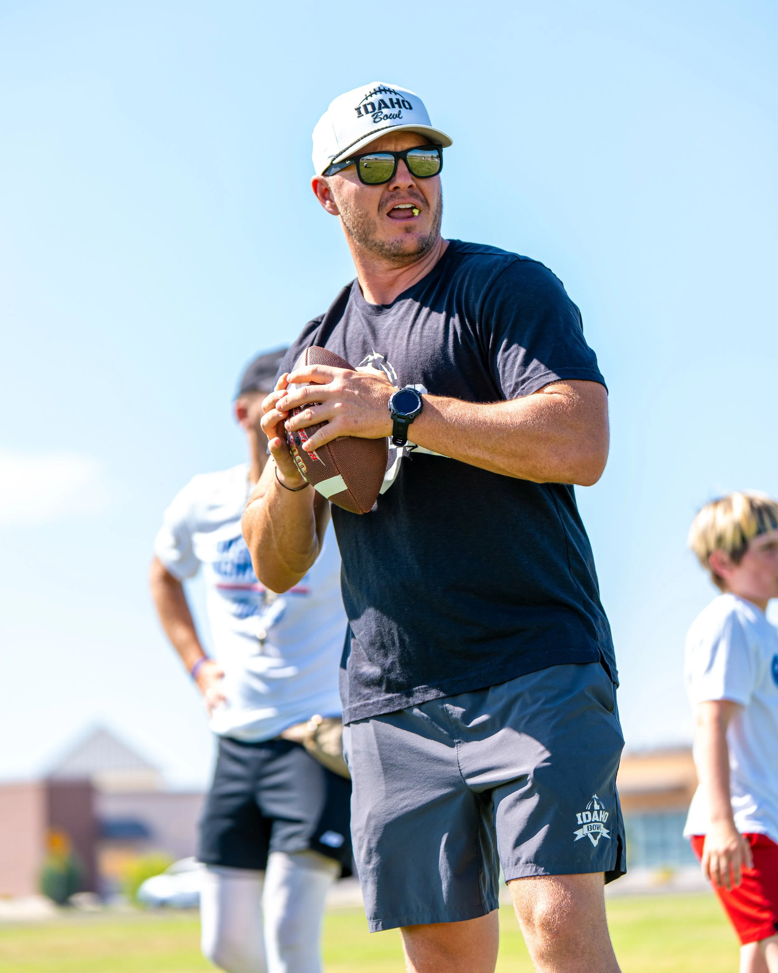A man wearing sunglasses, a white cap with 'Idaho Bowl' on it, a black t-shirt, and gray shorts holding a football during an outdoor sporting event.