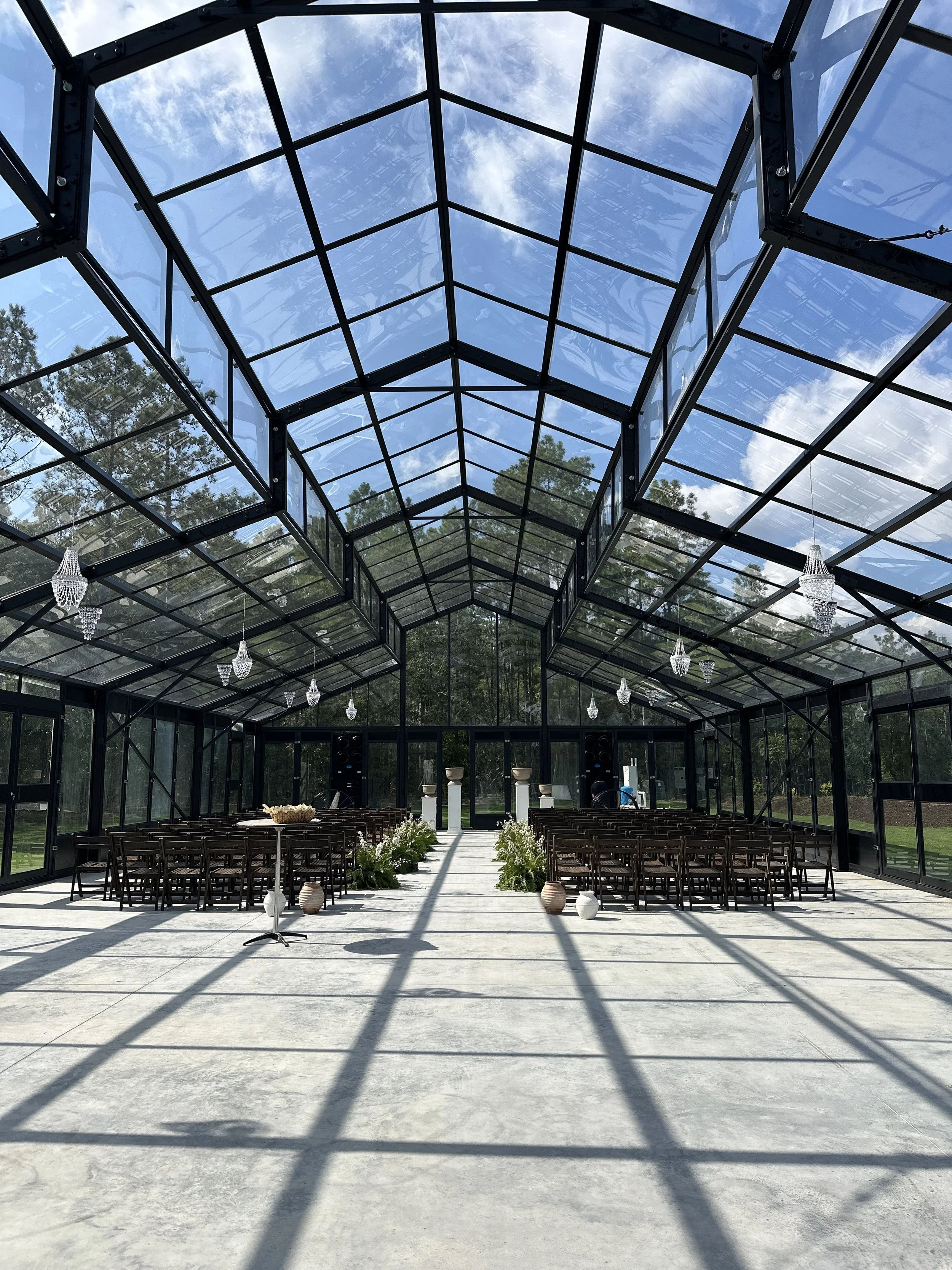 A modern glass building with black geometric framework, decorated for a wedding with pink and white flowers, fairy lights, and candle lanterns outside, during dusk.