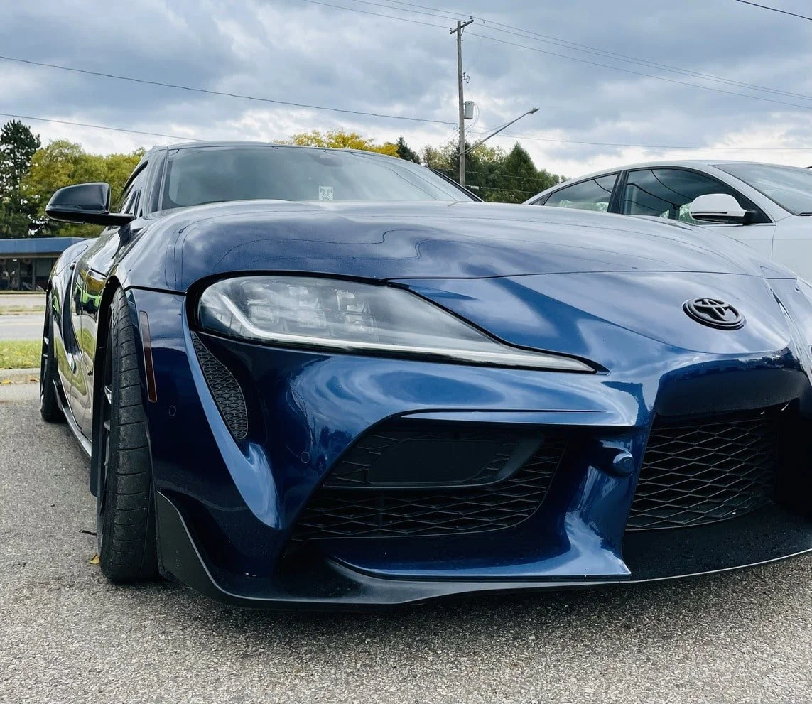 A blue Toyota sports car parked in a parking lot on a cloudy day, with another white car visible in the background.