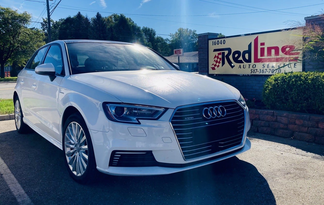 White Audi sedan parked outside Redline Auto Service near a sign with the business name and contact information.