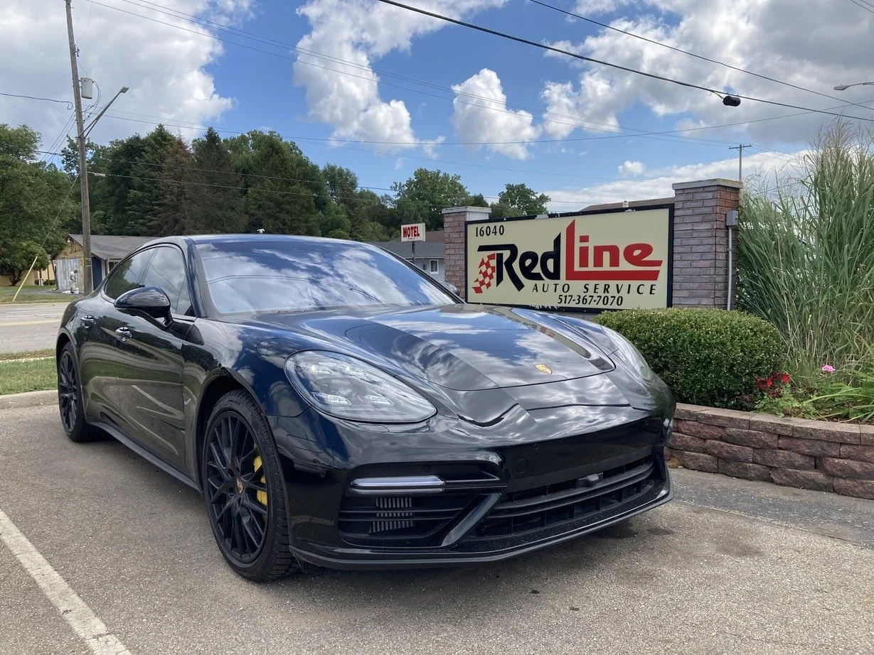 Black Porsche sports car parked in front of an auto service shop with a sign reading Red Line Auto Service, under a cloudy sky.