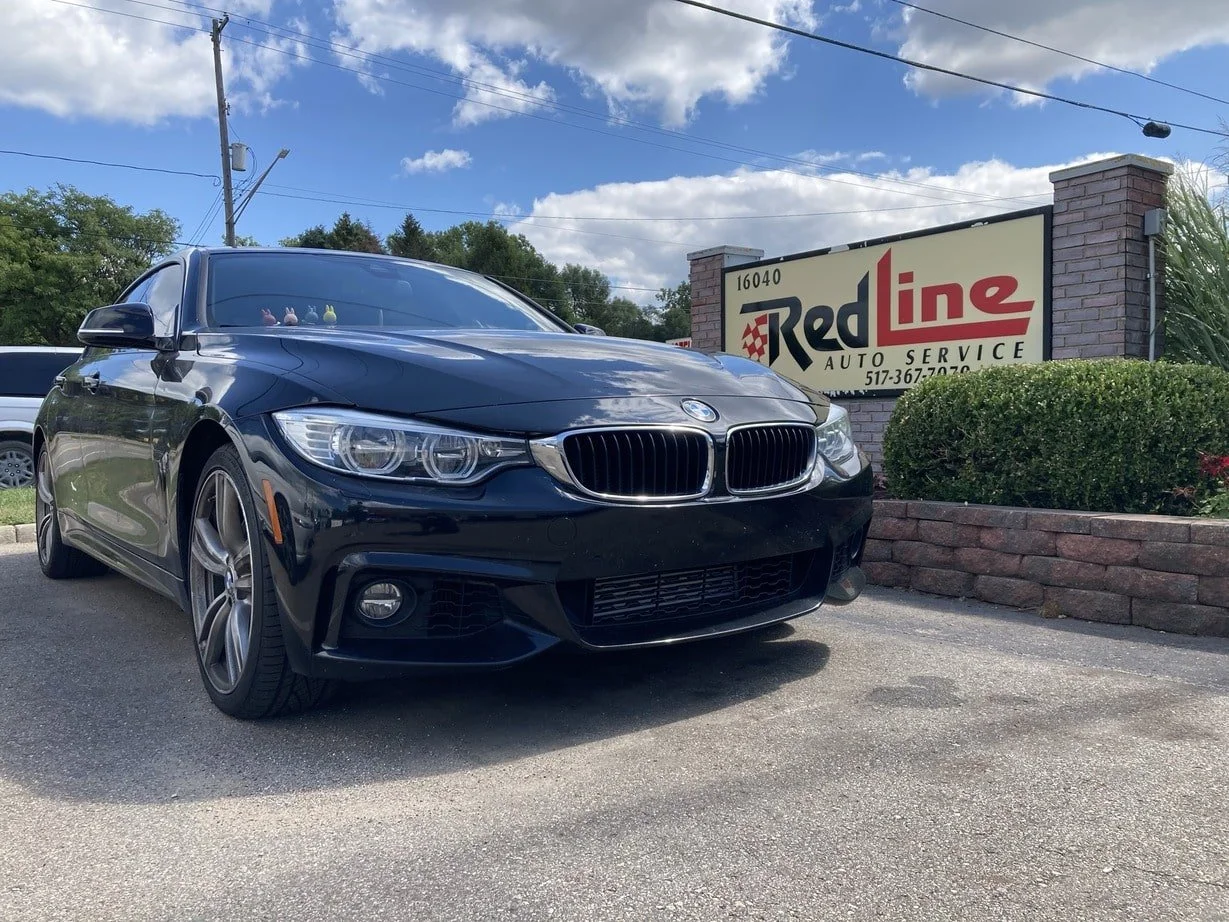 Black BMW sedan parked outside RedLine Auto Service with a brick sign and bushes, under partly cloudy sky