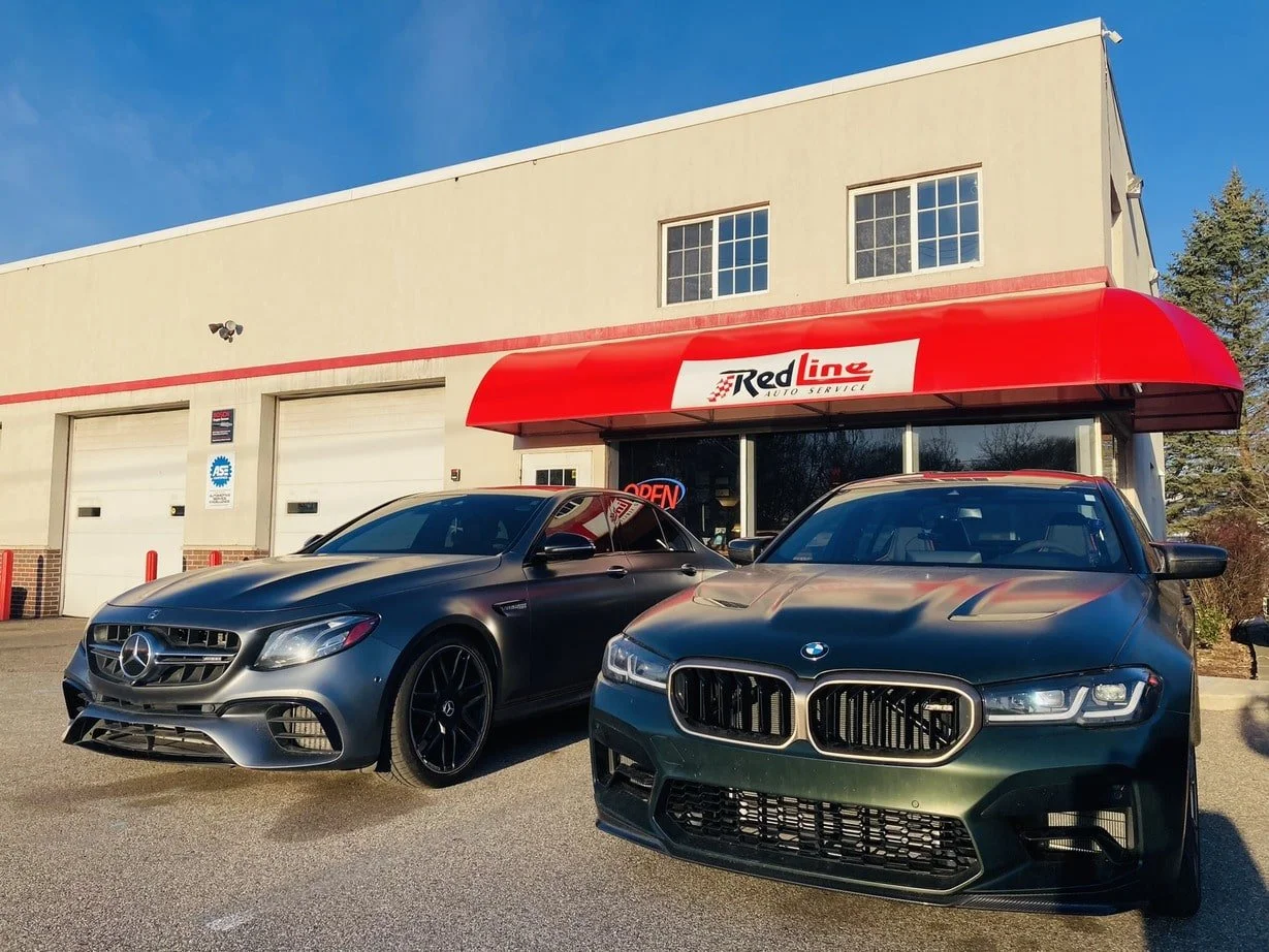 A dealership with a red awning labeled 'Red Line Auto Service', two luxury cars - a gray Mercedes-Benz and a black BMW parked in front, and a warehouse-style building with garage doors behind them.