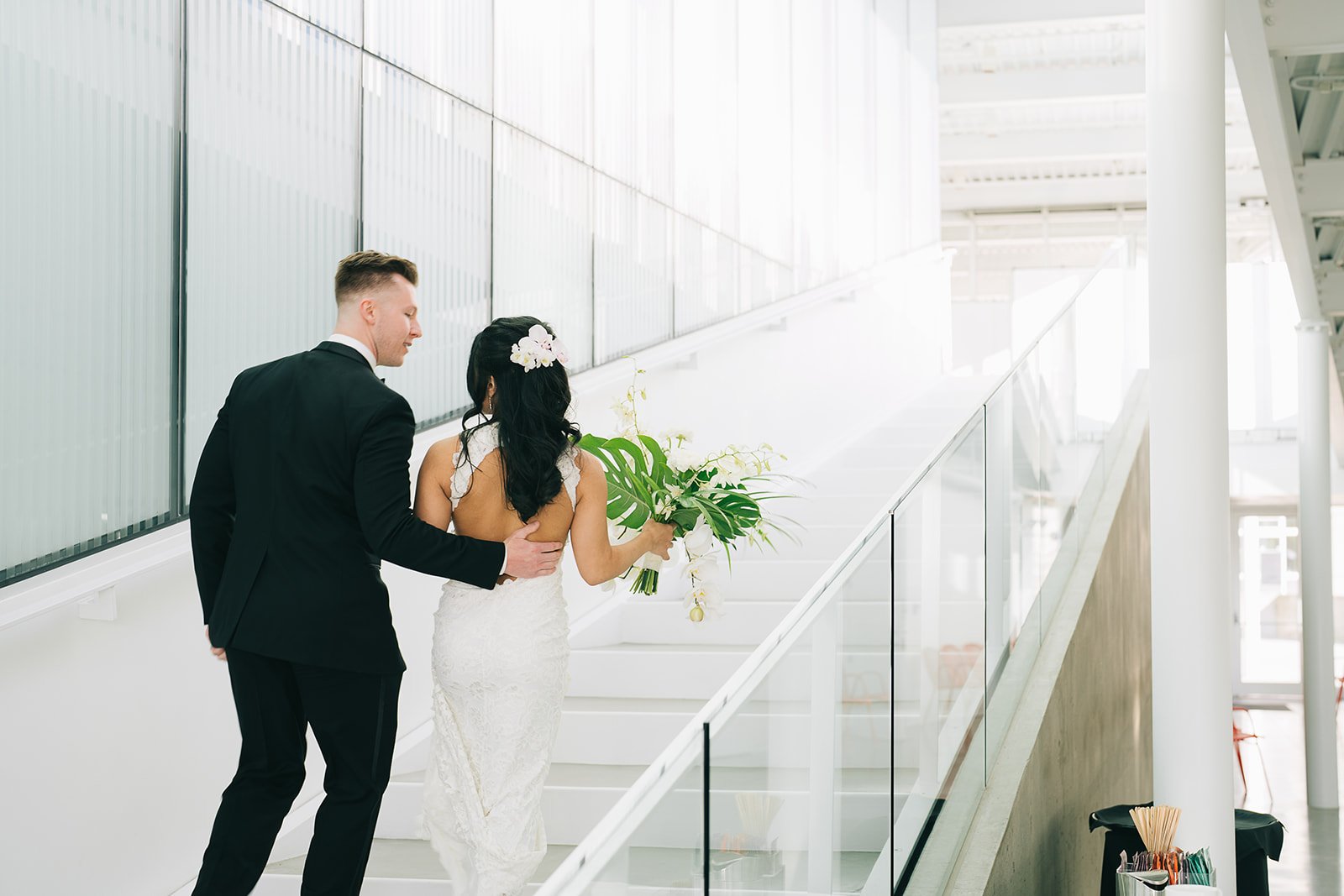 Bride and groom ascending modern staircase, bride holding bouquet, back view.