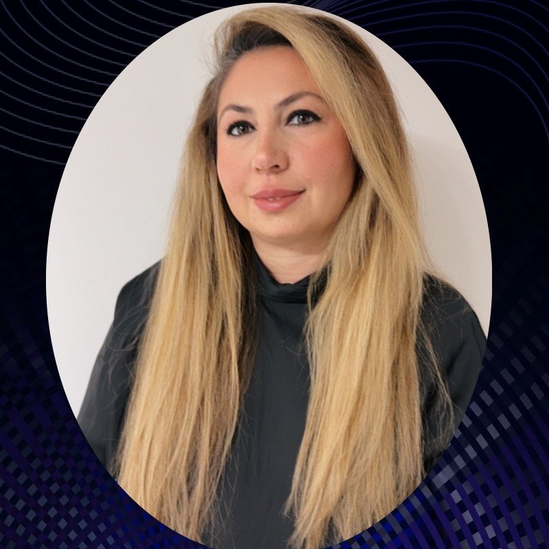 A woman with long blonde hair and light makeup, wearing a black top, posing against a plain white background.