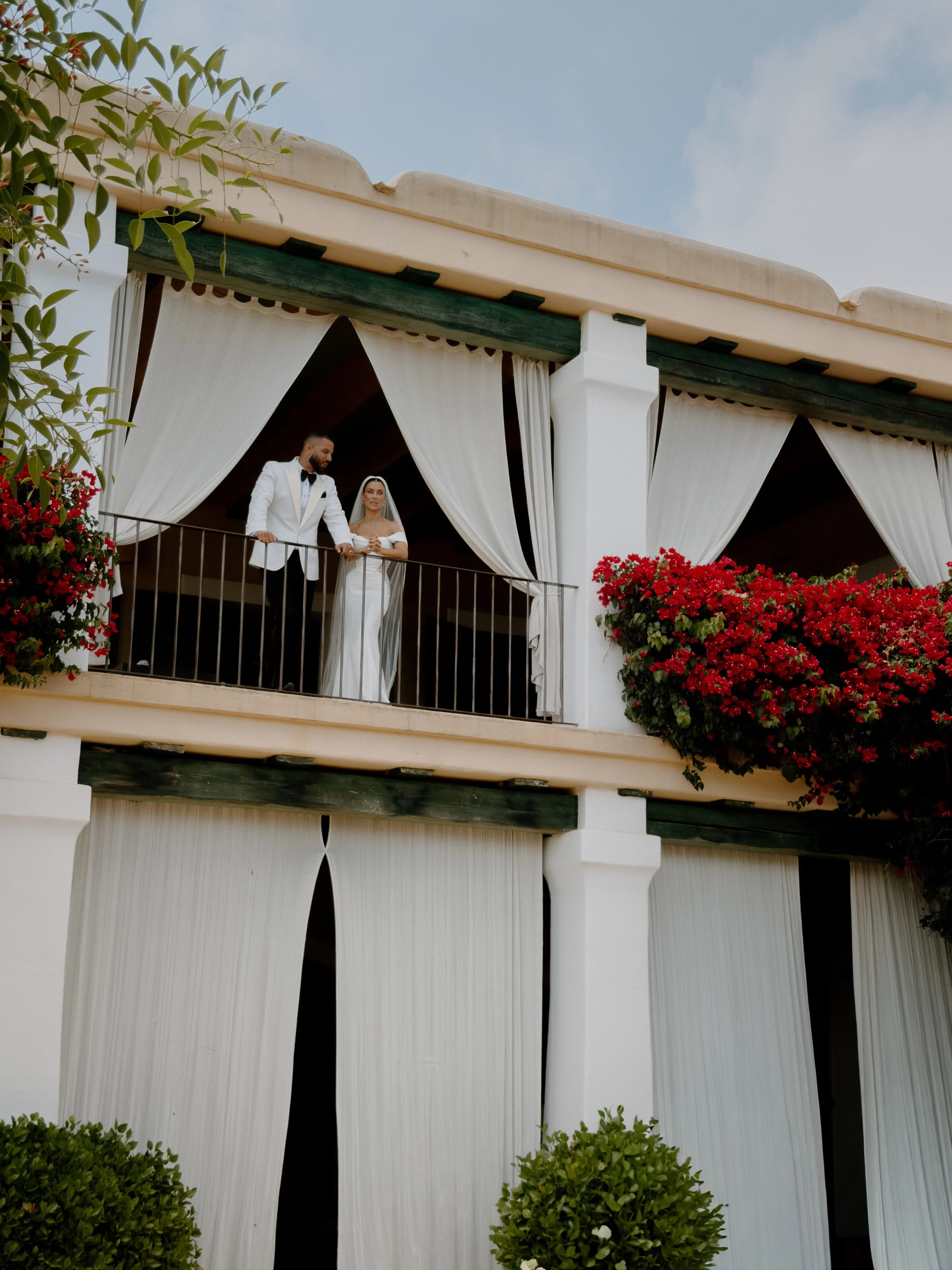 A bride and groom in wedding attire standing on a balcony with white curtains and red flowers, overlooking a clear sky.