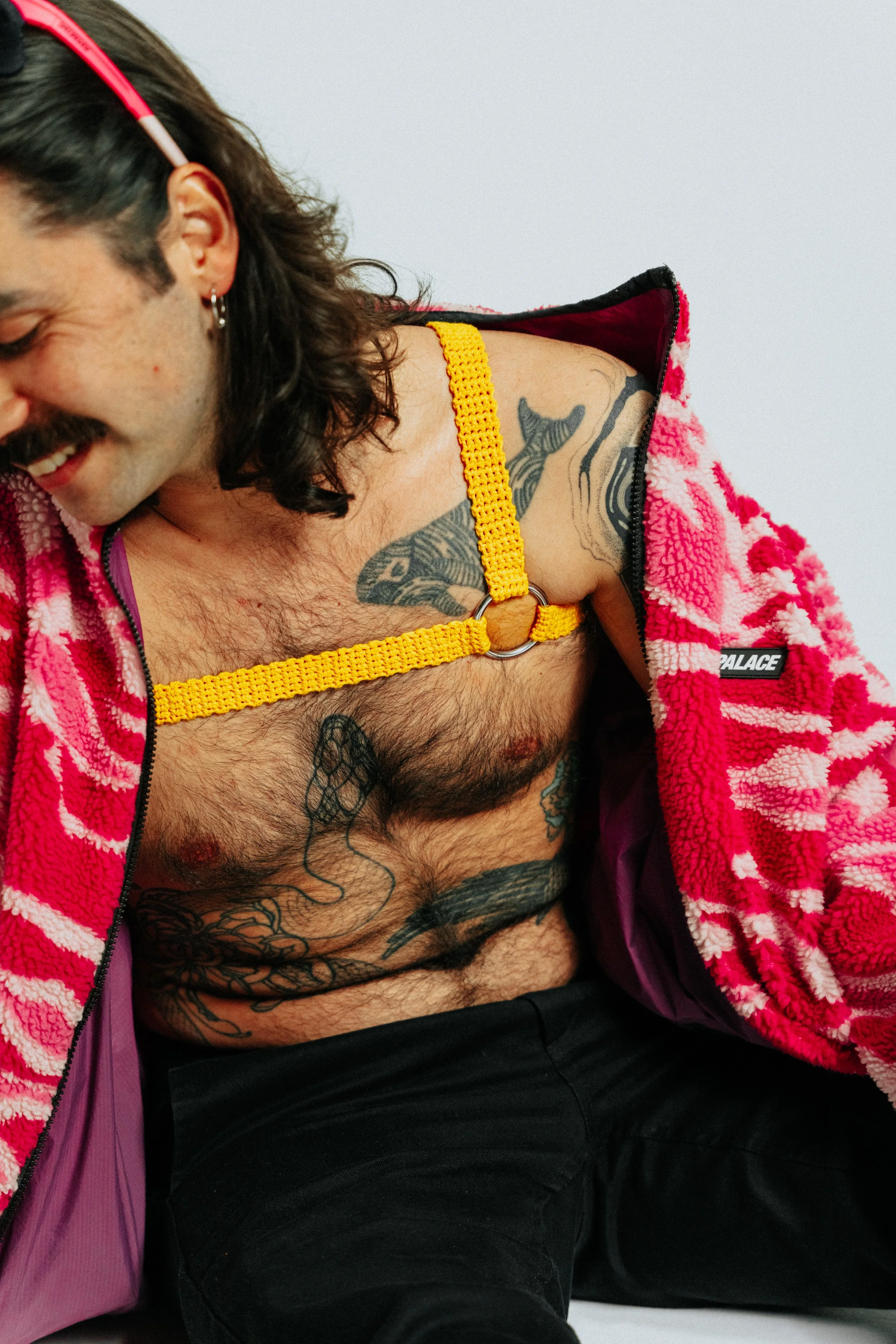 A smiling man with long dark hair, earrings, and tattoos on his chest and torso, wearing a pink and red fuzzy jacket and a yellow harness.