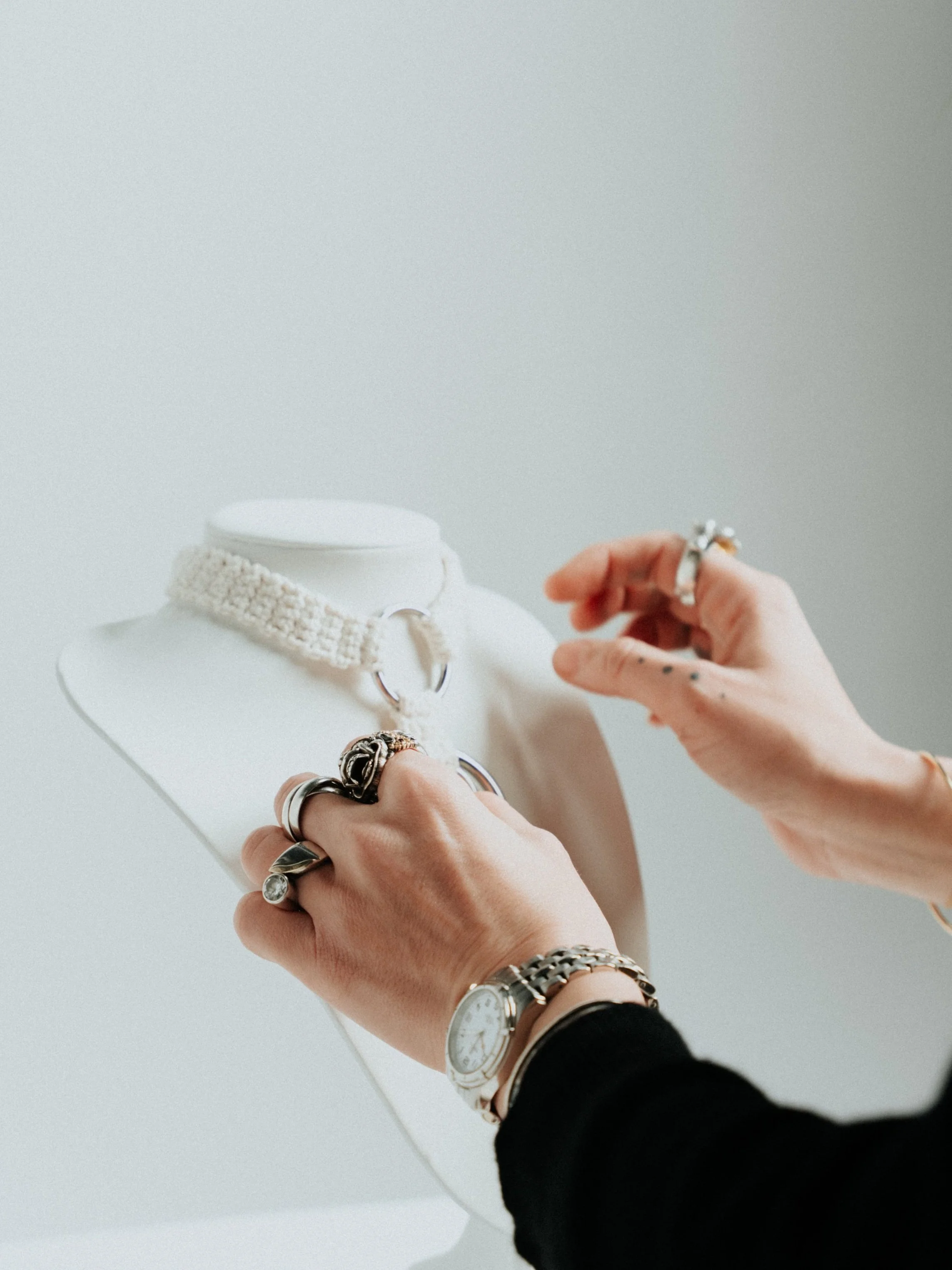 Person adjusting jewelry on a jewelry display bust, wearing multiple rings and a watch, with a choker necklace on the bust.