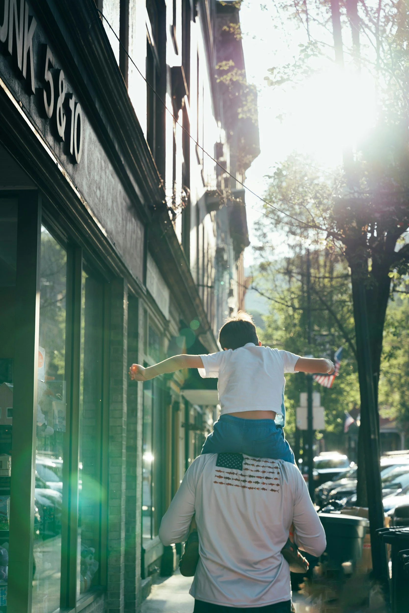 A child riding on an adult's shoulders on a sidewalk in a sunlit street with trees and buildings.