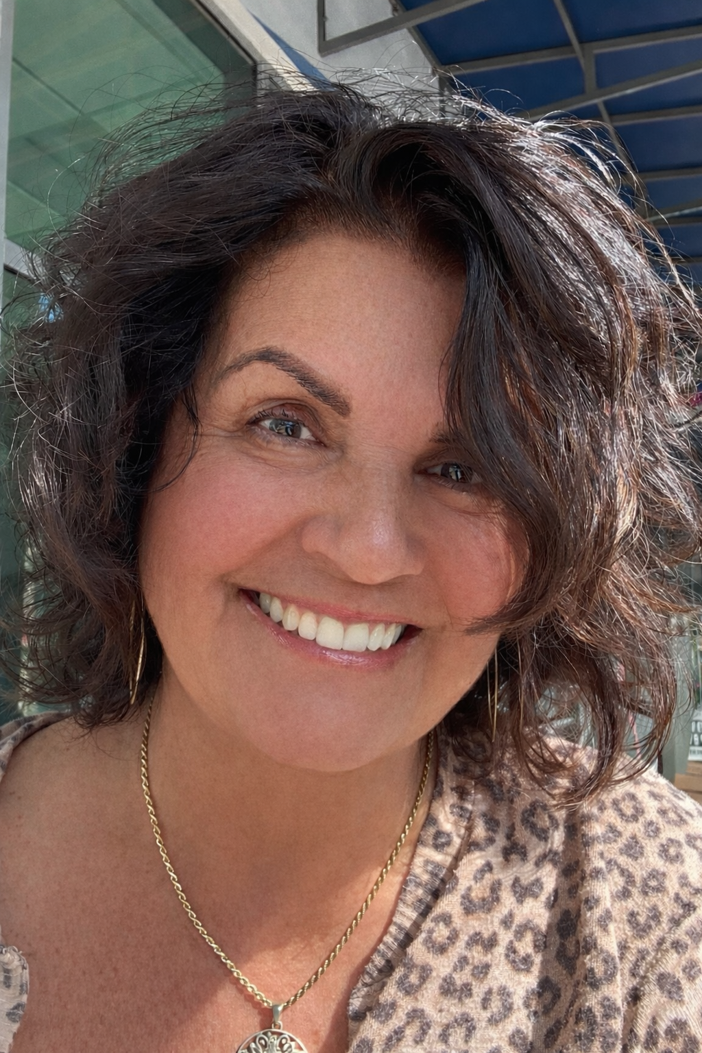 Close-up of a smiling woman with curly dark brown hair, wearing a leopard print top, gold necklace, and hoop earrings, outdoors with a building and railing in the background.