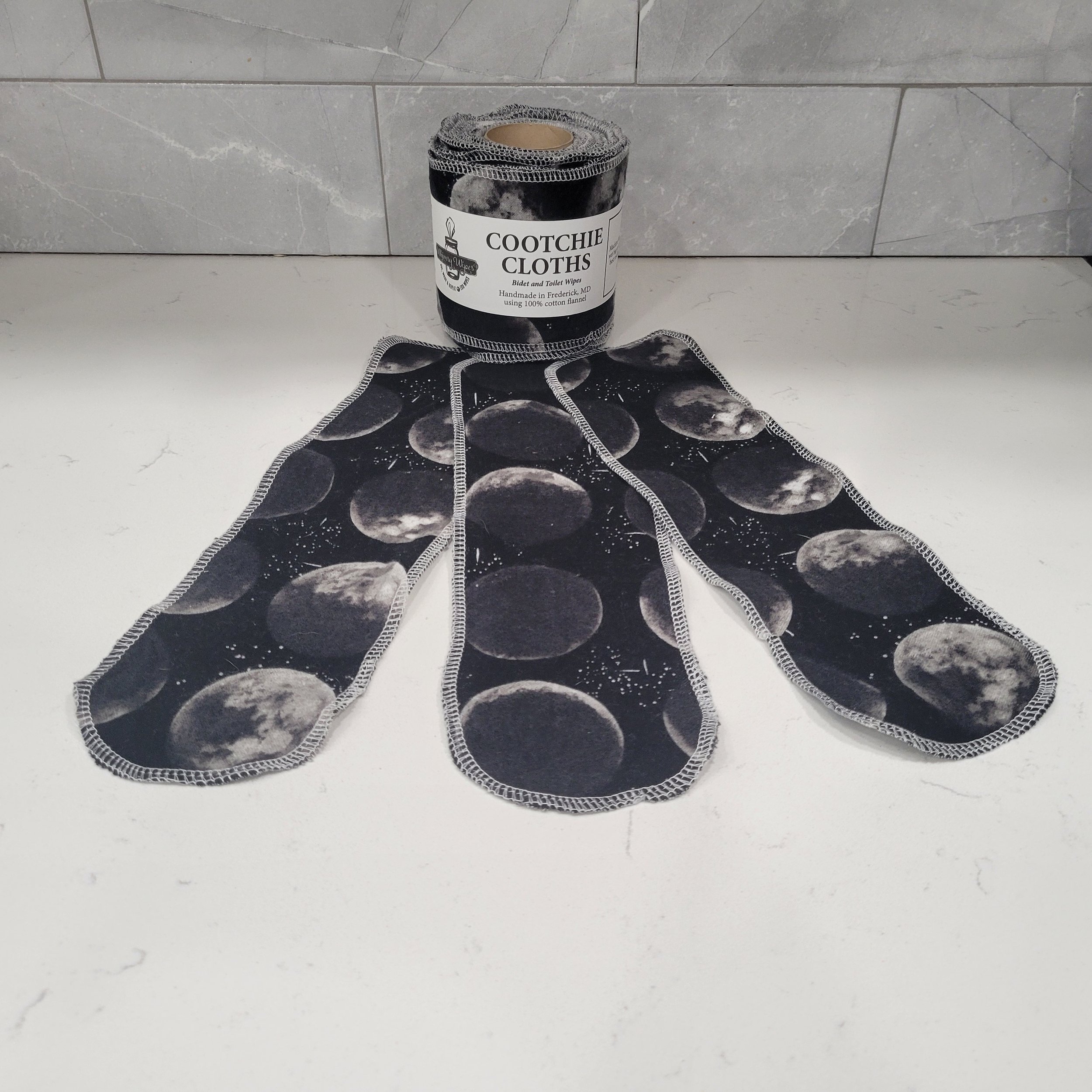 A single roll of reusable toilet paper with three strips in front to display the 12 inch length. Each cloth is showing the phases of the moon on a black background
