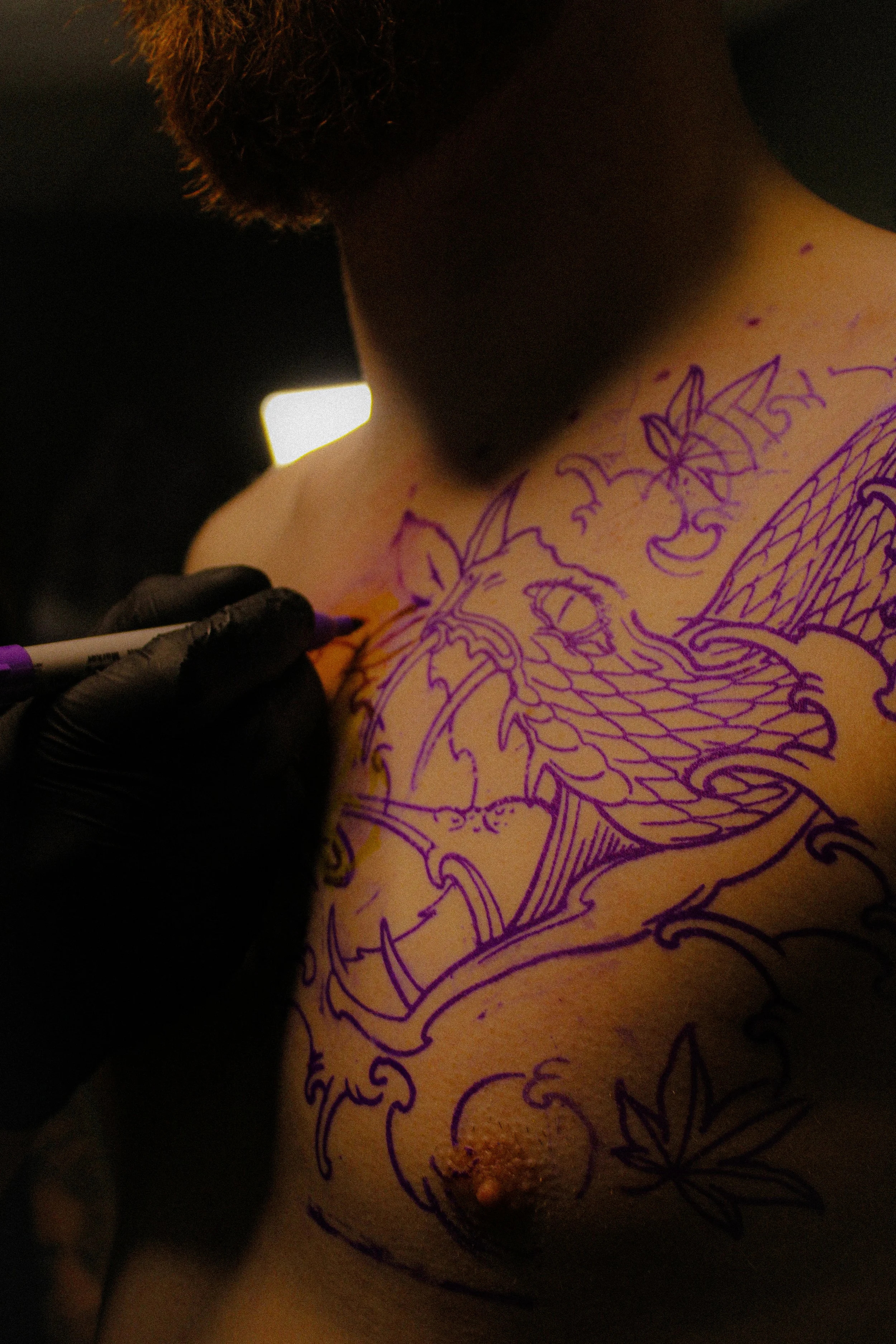 A person with a beard is getting a tattoo on their chest. The tattoo design is made with purple ink and features intricate floral and mystical elements.