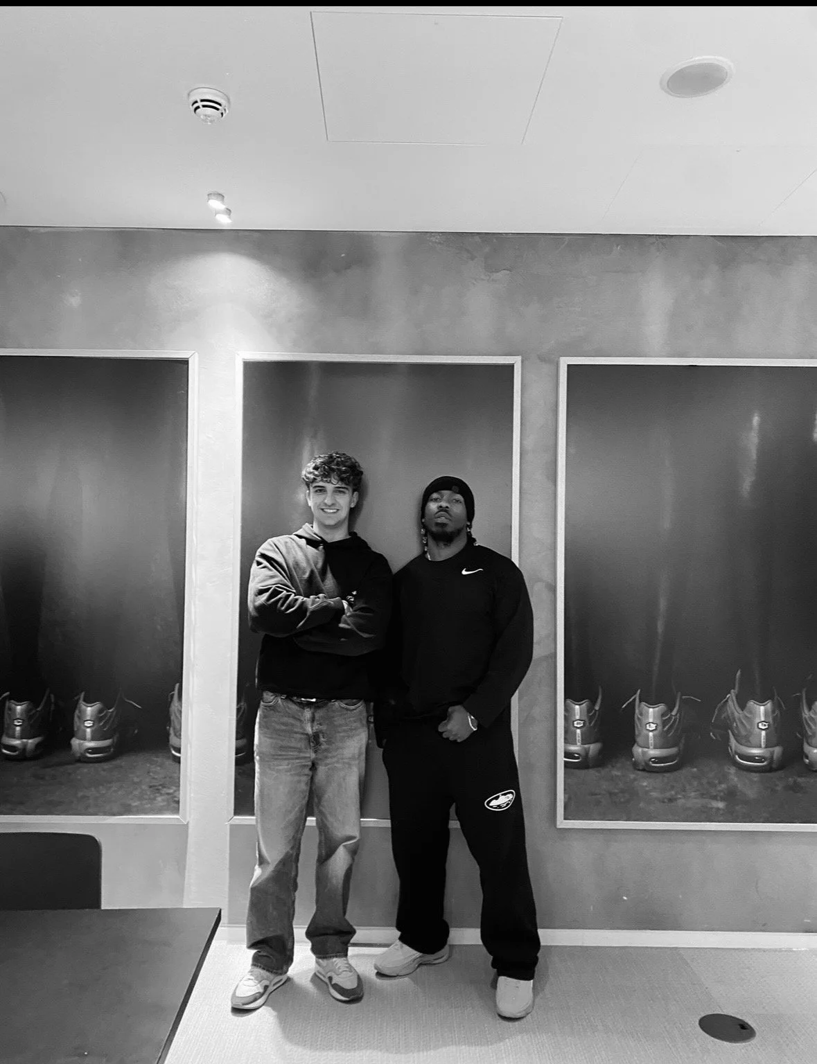 Two young men standing next to each other, posing for a photo in front of a wall with display cases containing sneakers. One is smiling with arms crossed, the other has a serious expression, wearing a beanie and Nike sweat suit.