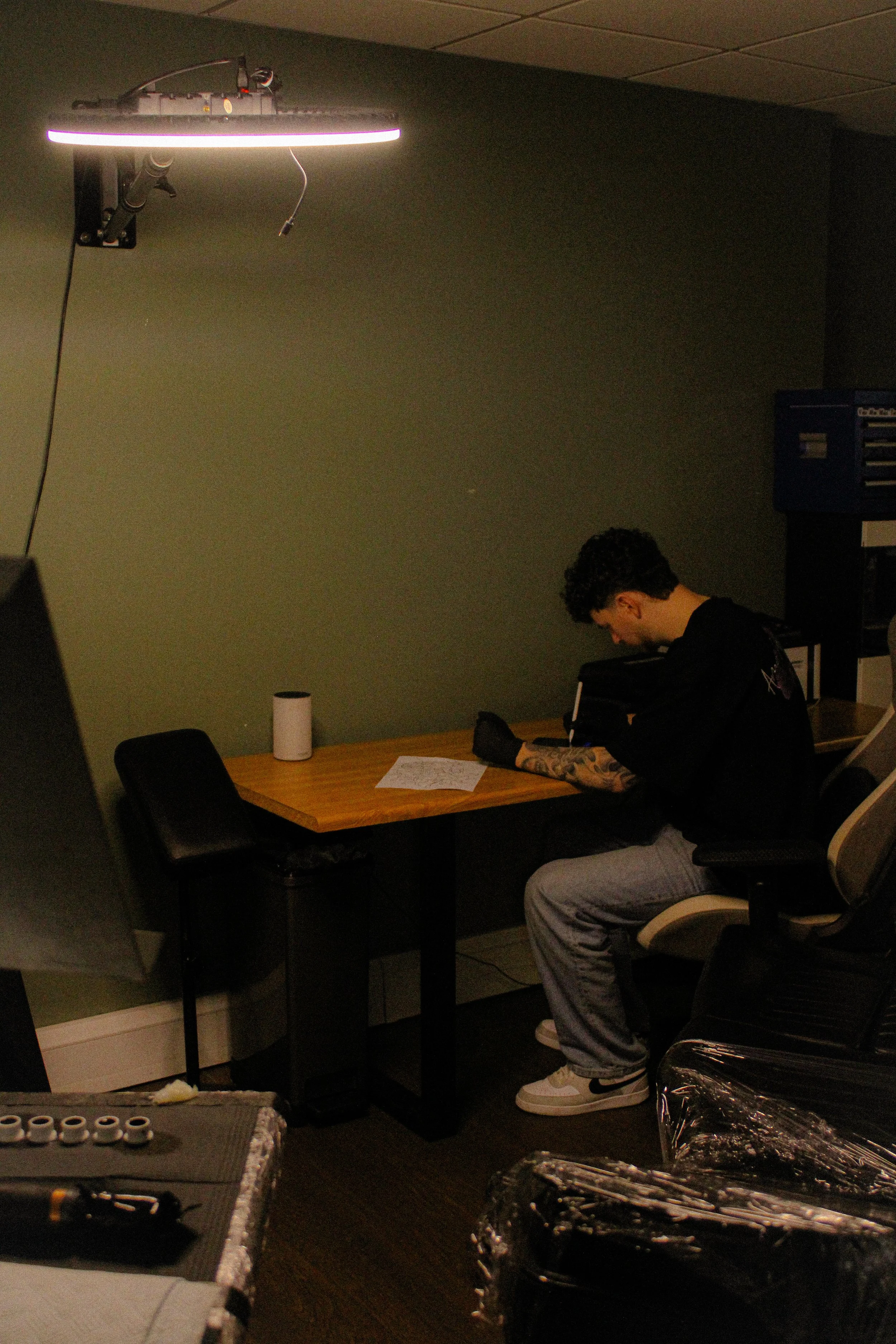 A person with tattoos on their arms sitting at a desk in a dark room, looking down at their phone. In the background, there are equipment and items on tables, and a large illuminated ring light on the ceiling.