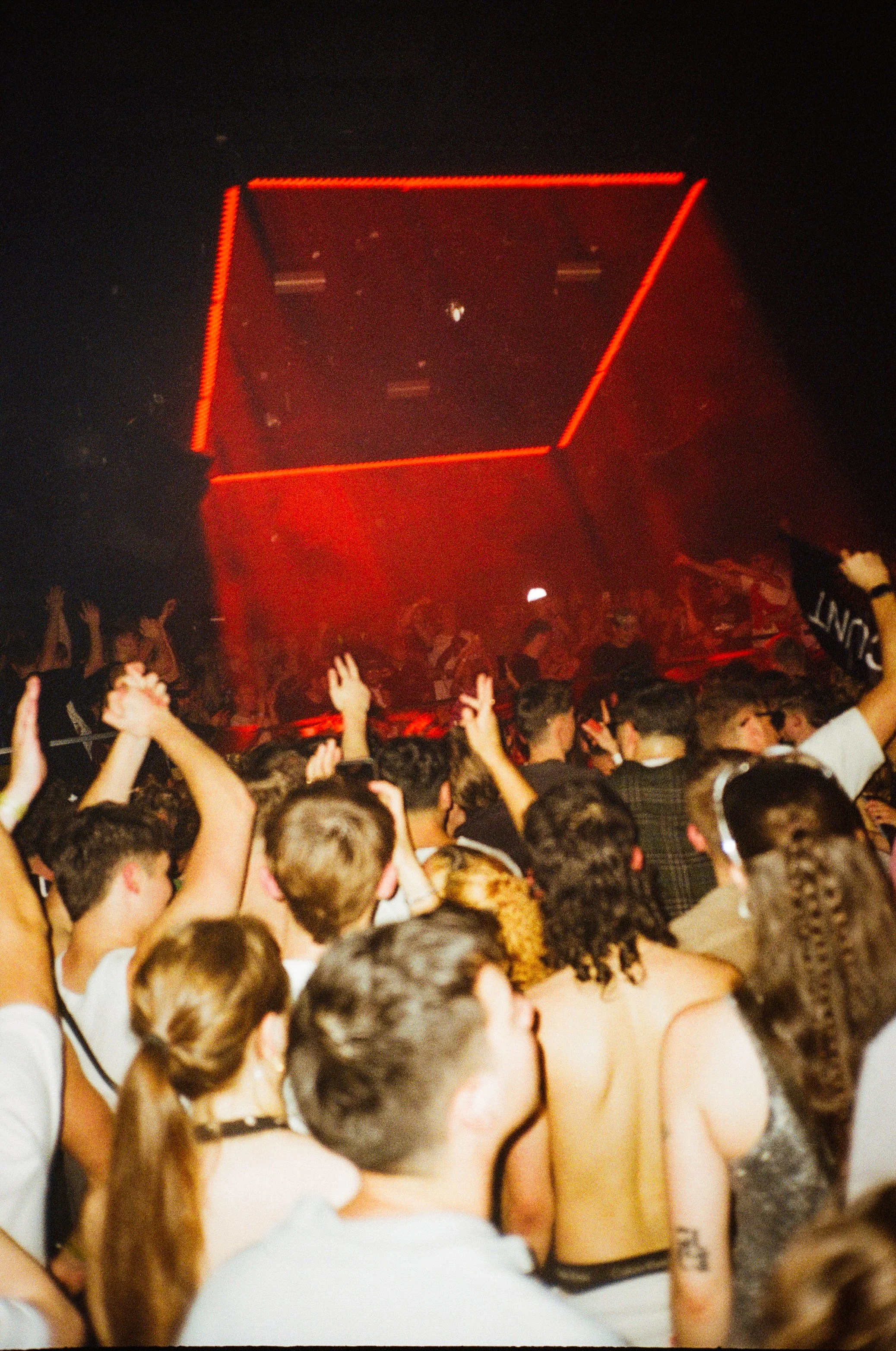 A crowded dance floor with many young people dancing and raising their hands, illuminated by red lighting, at a party or club.