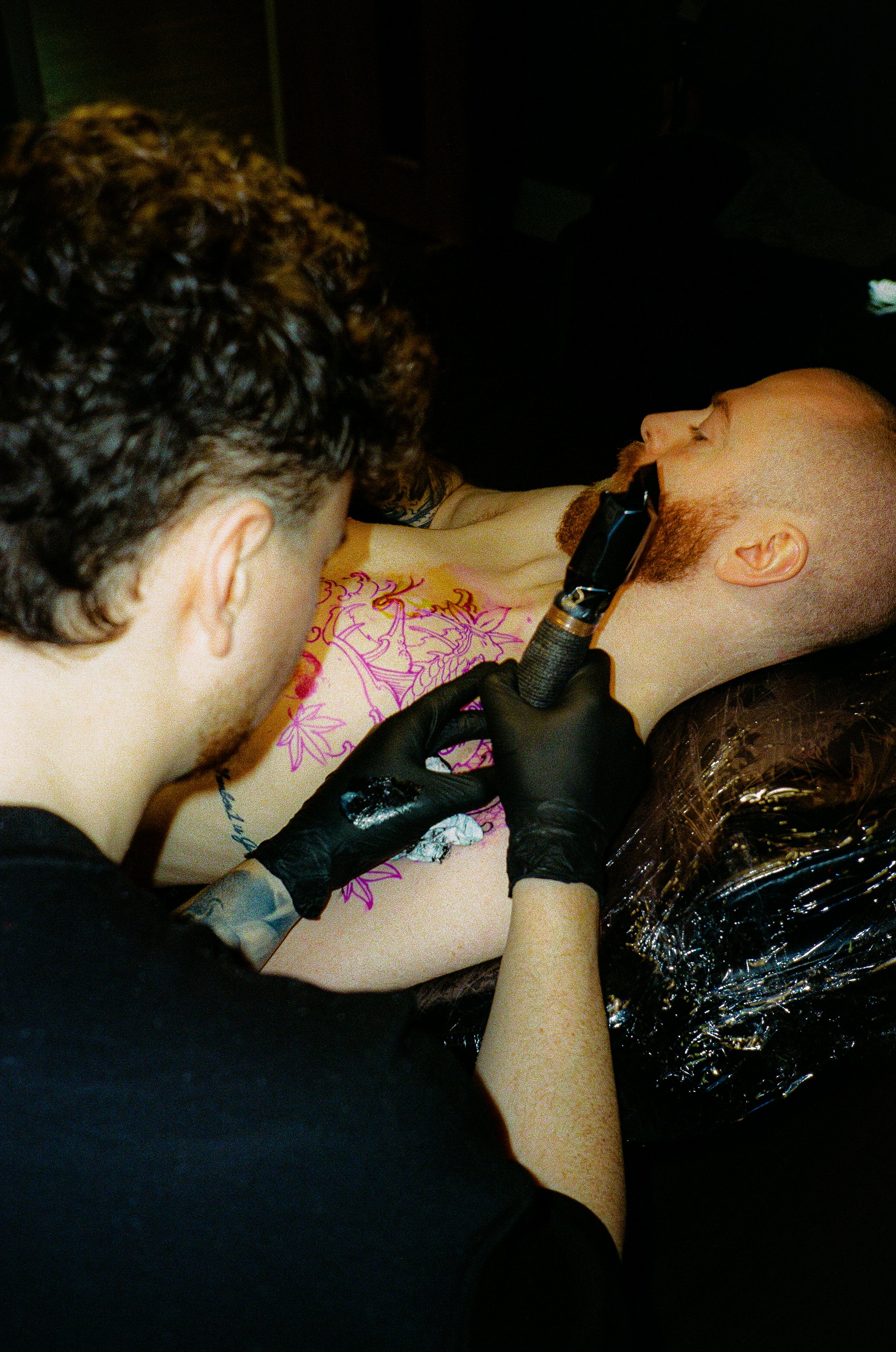 A person with black gloves is using a tattoo machine on a man's neck, who has his eyes closed and is lying on a tattoo table covered with plastic wrap, in a dimly lit setting.