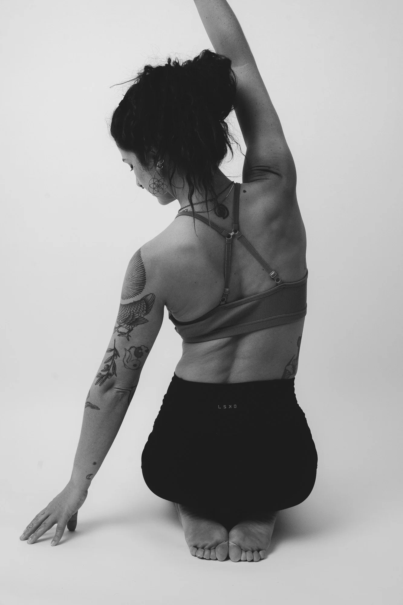 Black and white photo of a woman doing a yoga pose with tattoos on her arm, wearing a sports bra and leggings, kneeling on the floor with one arm extended upward.