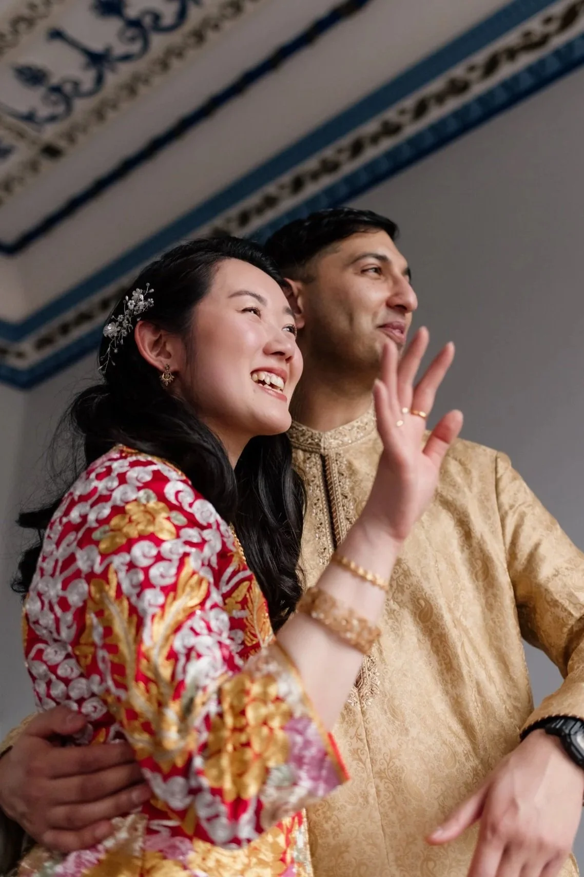 A smiling couple dressed in traditional attire, the woman in a red and gold embroidered dress and the man in a gold embroidered shirt, waving and posing together indoors.