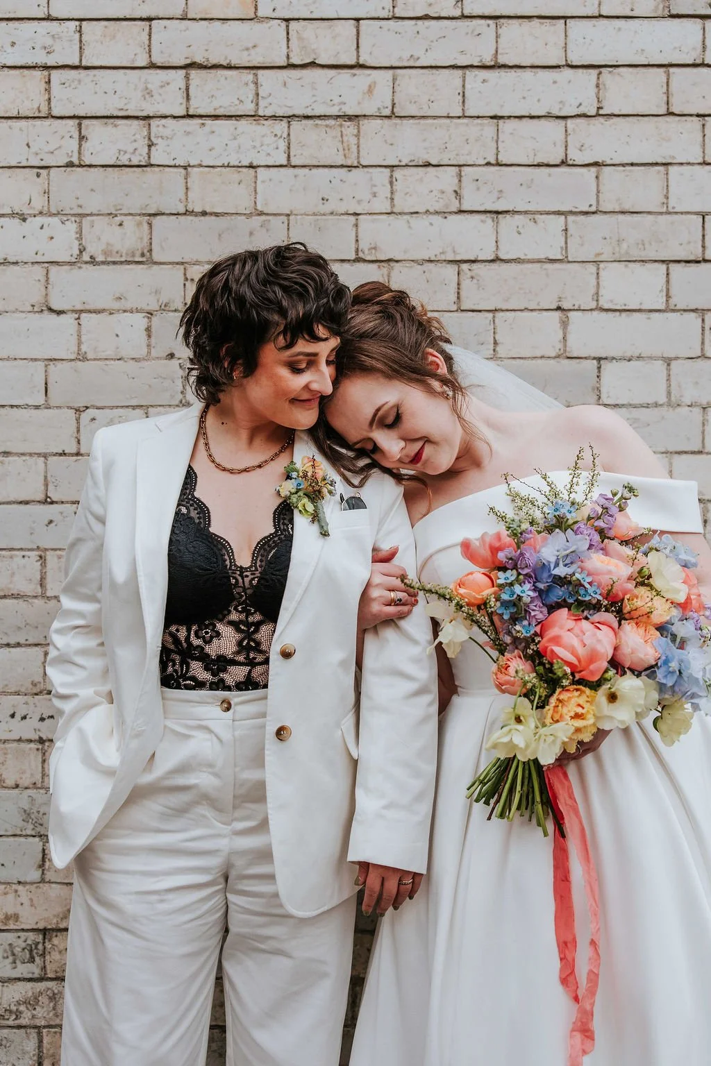 Two women, one in a white suit with black lace top and the other in a white wedding dress holding a colorful bouquet, leaning their heads together against a brick wall.