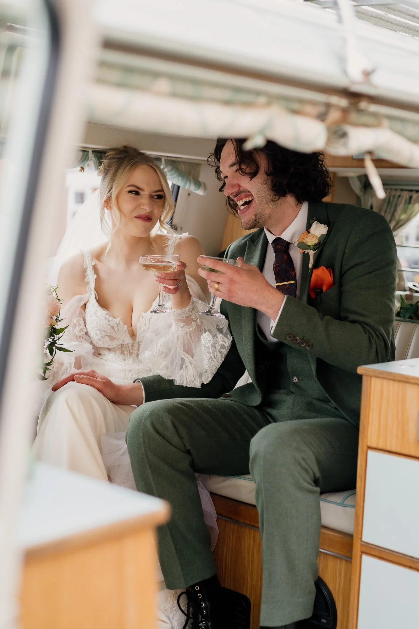 A bride and groom sitting in a vintage trailer, enjoying drinks and sharing a playful moment. The bride wears a lace wedding dress, and the groom is in a green suit with a boutonnière.