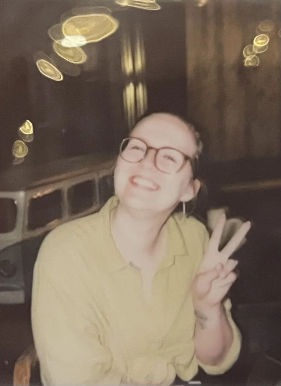 A woman with glasses and a yellow shirt smiling and making a peace sign in a dimly lit setting with blurred bokeh lights.