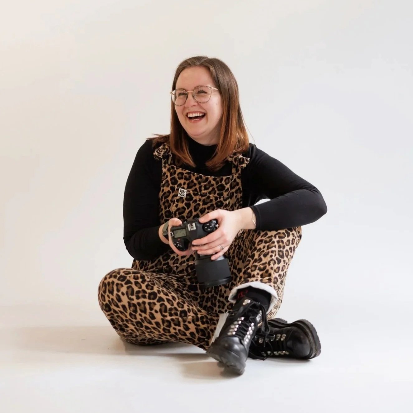 A woman sitting cross-legged on the floor, smiling and holding a camera, wearing a leopard print jumpsuit, black long-sleeve shirt, black boots, and glasses, against a plain white background.