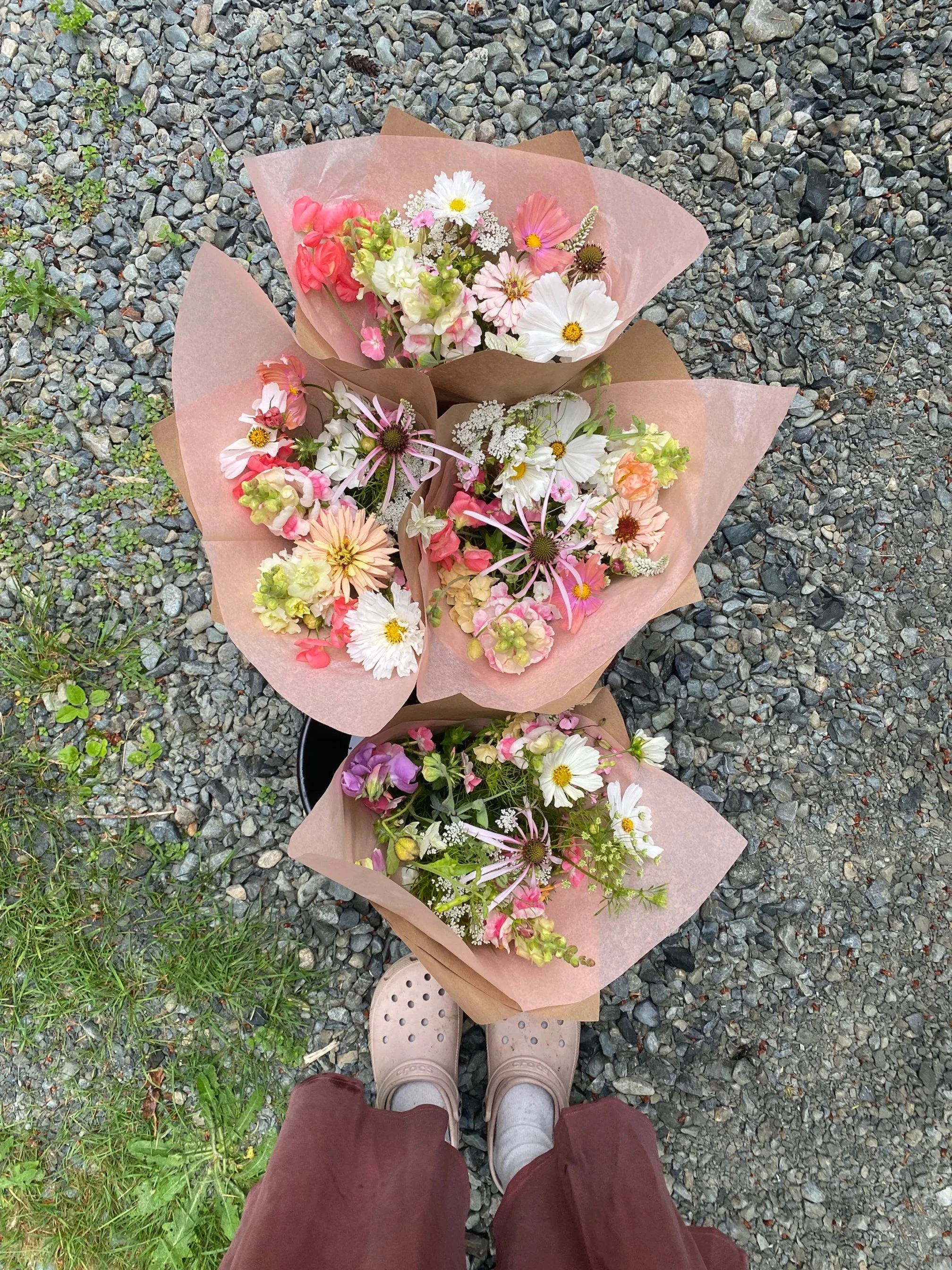 Three bouquets of mixed flowers including daisies, and other small flowers, wrapped in pink and brown paper, viewed from above, with someone's feet in sandals and maroon pants visible.