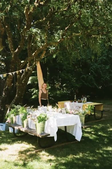 Outdoor scene with a woman standing near a table decorated with flowers and drinks, under a large tree with green foliage.
