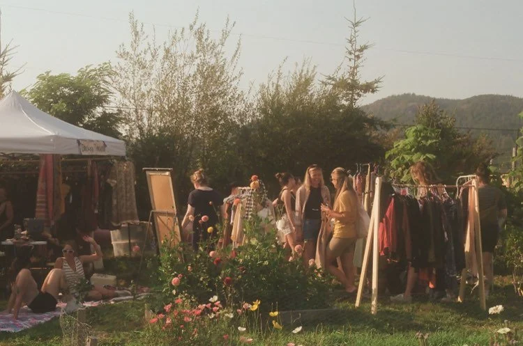 People browsing clothing and accessories at an outdoor market on a sunny day, with trees, flowers, and mountains in the background.
